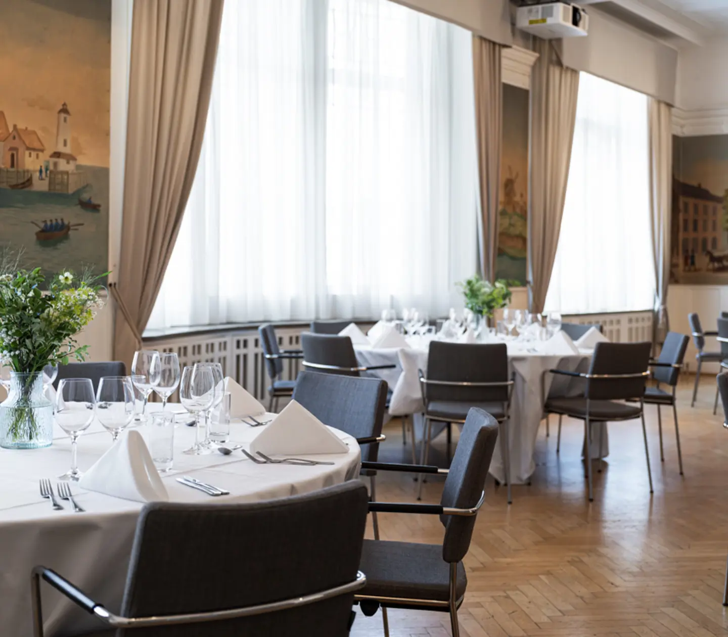 Elegant middagsmiljö med dukade runda bord, blommor i vaser, och smakfull inredning i en ljus sal med stora fönster och målade väggar.