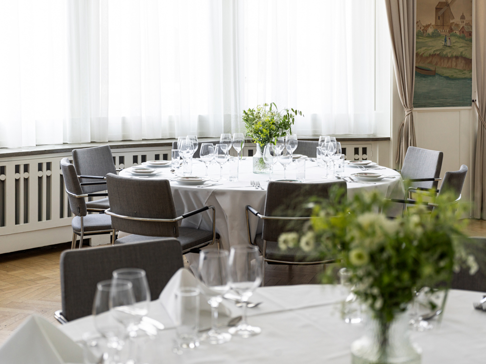 Elegant restauranginteriör med dukade runda bord, vita dukar, vinglas och blomsterarrangemang i ljus, luftig miljö.