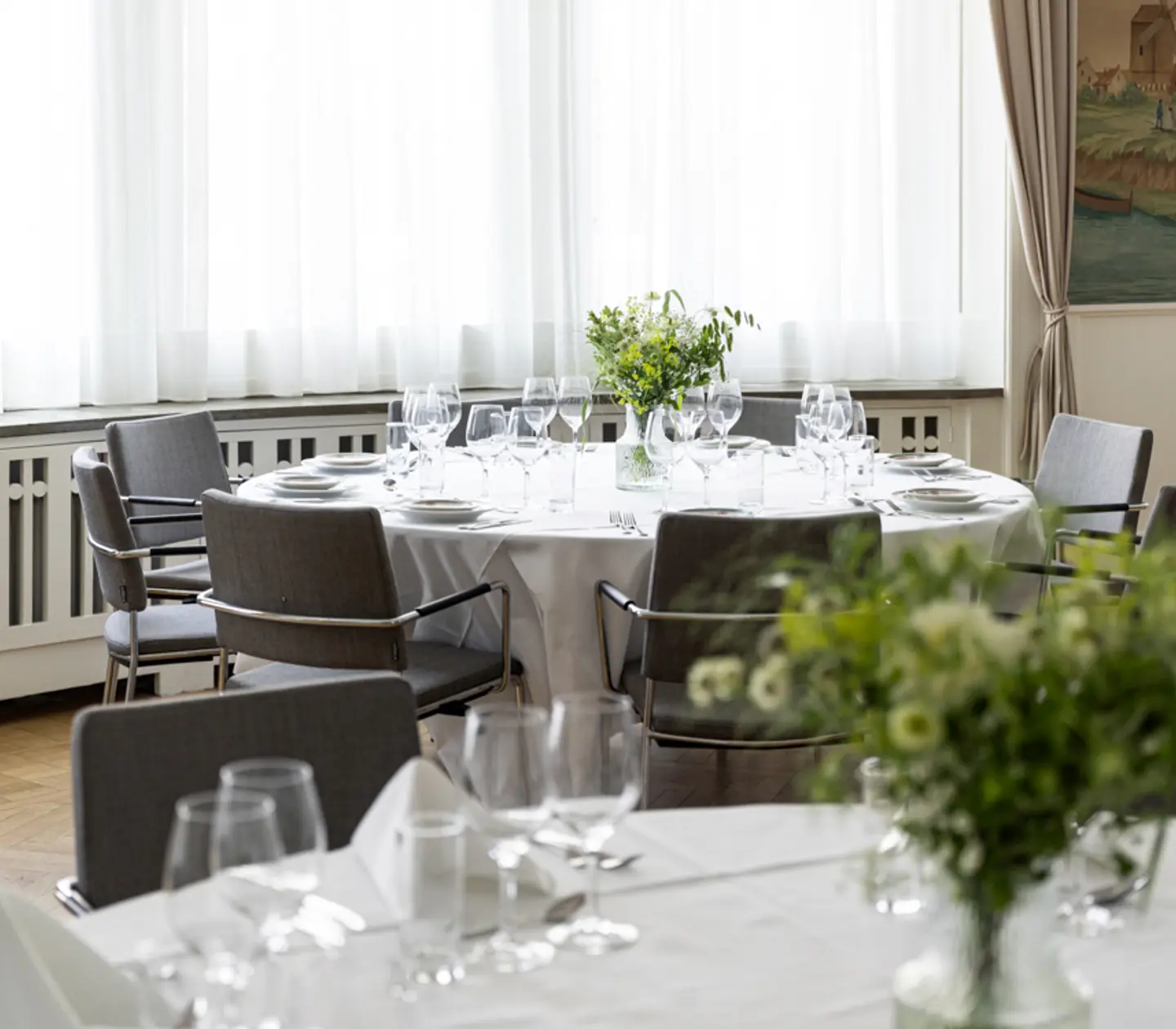 Elegant restauranginteriör med dukade runda bord, vita dukar, vinglas och blomsterarrangemang i ljus, luftig miljö.