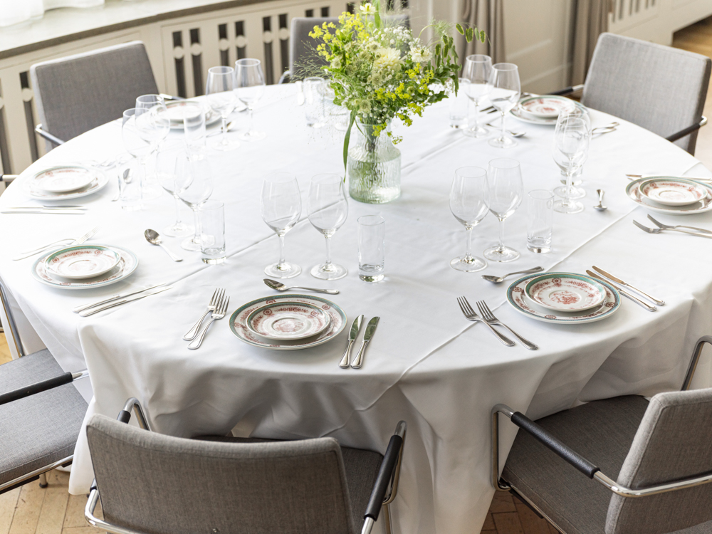 Rundt bord dukat för middag med vit duk, porslinstallrikar, bestick och vinglas, samt en vas med gröna blommor i mitten.
