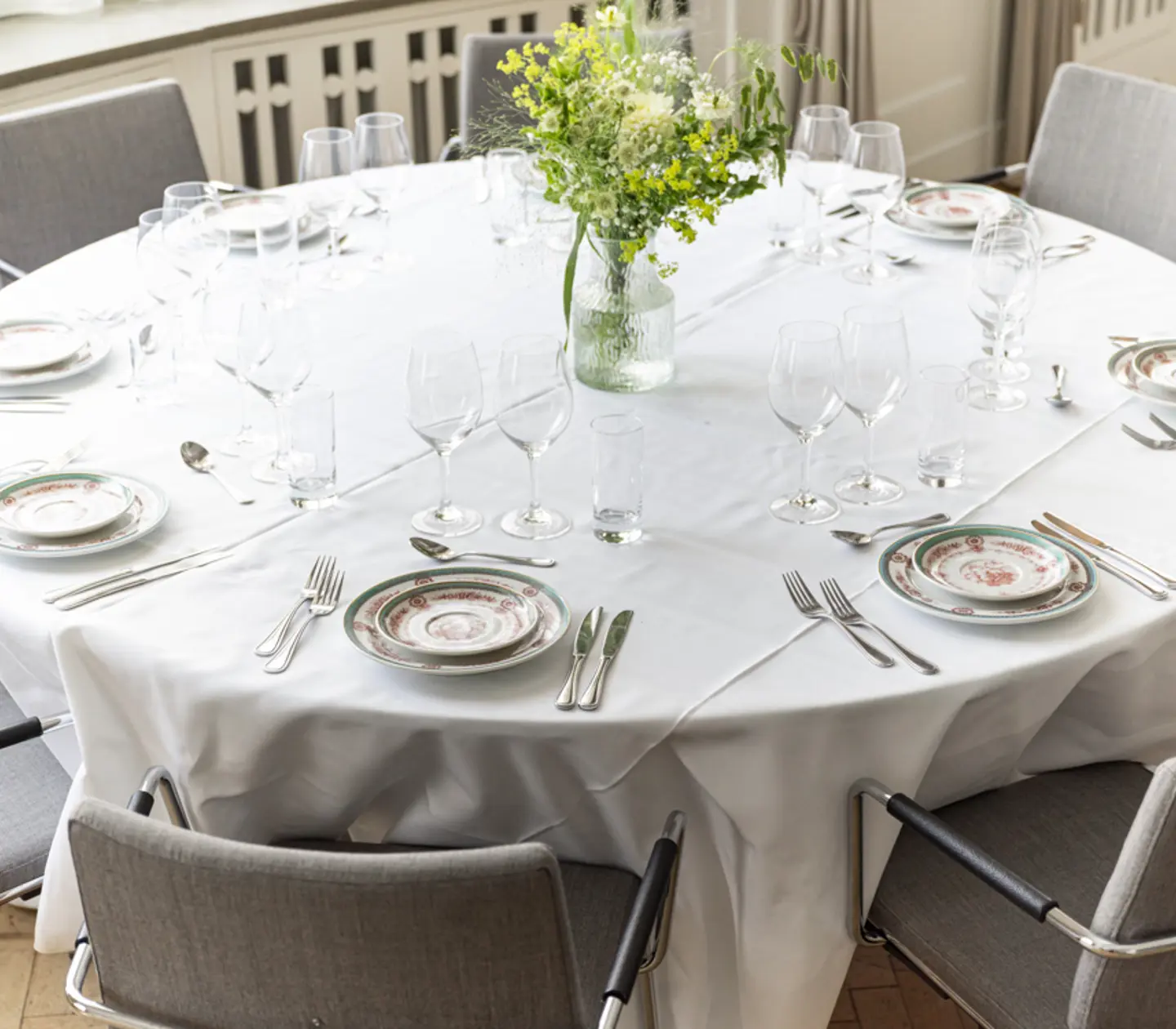 Rundt bord dukat för middag med vit duk, porslinstallrikar, bestick och vinglas, samt en vas med gröna blommor i mitten.