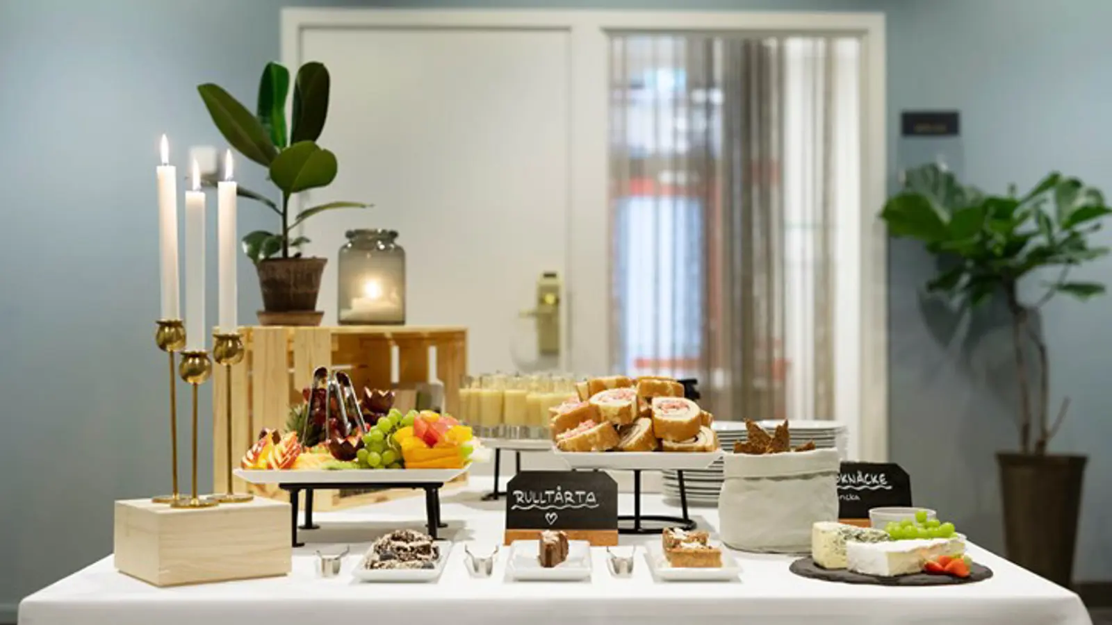 Närbild på buffébord med vit duk, fika, frukt, ostar och tända ljus