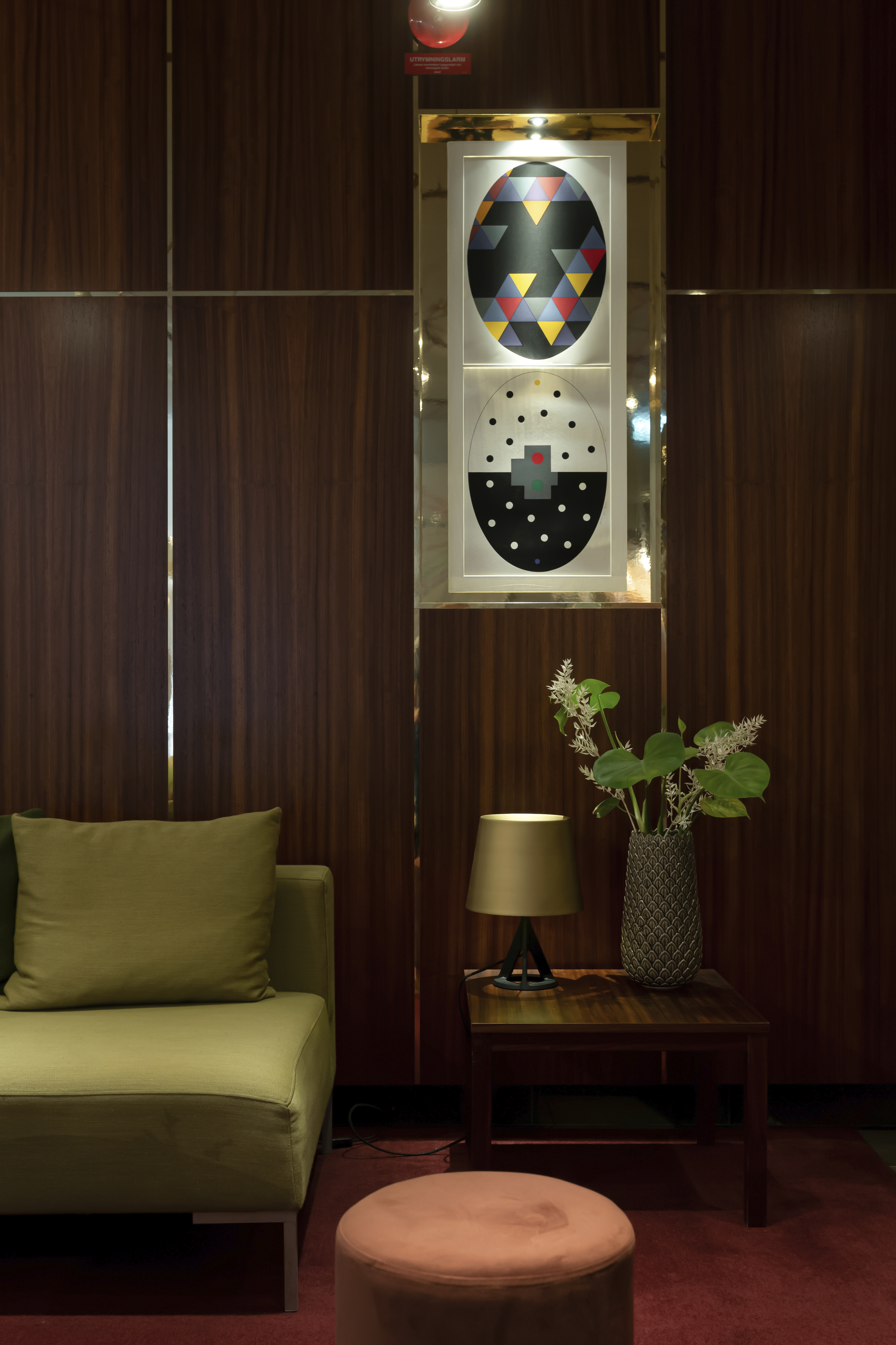 Modern interior with stylish olive green sofa, wooden wall paneling, and abstract geometric artwork. A side table with a decorative plant and lamp enhances the cozy, contemporary living space.