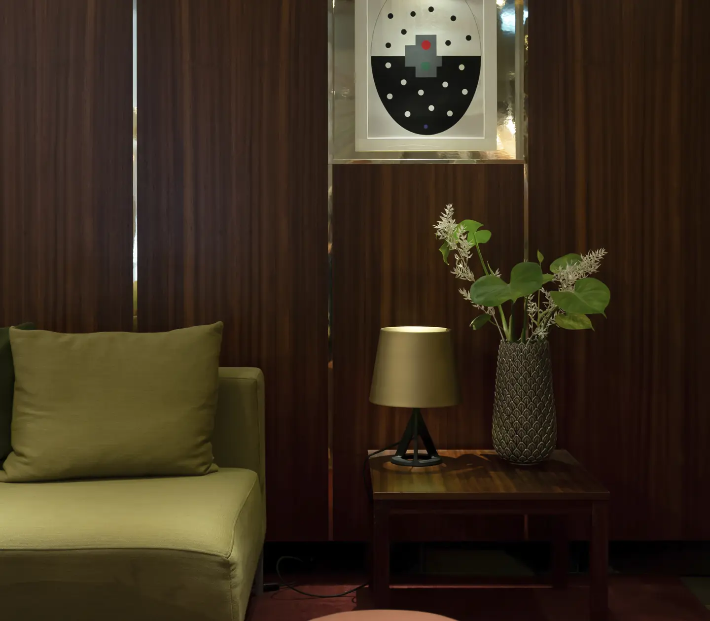 Modern interior with stylish olive green sofa, wooden wall paneling, and abstract geometric artwork. A side table with a decorative plant and lamp enhances the cozy, contemporary living space.