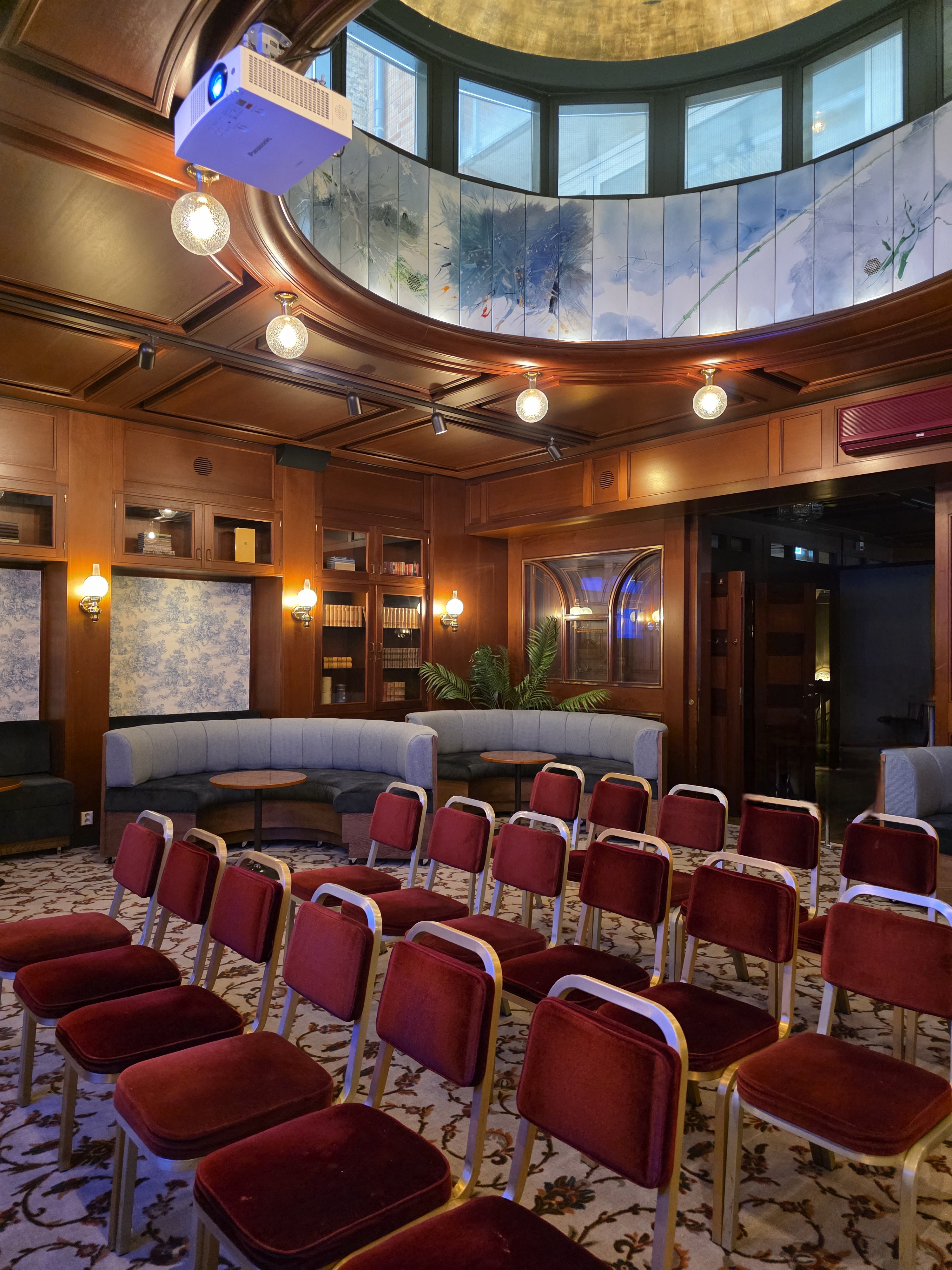 Elegant event space with red velvet chairs arranged in rows, cozy seating areas, wooden paneling, ambient lighting, and decorative ceiling details.