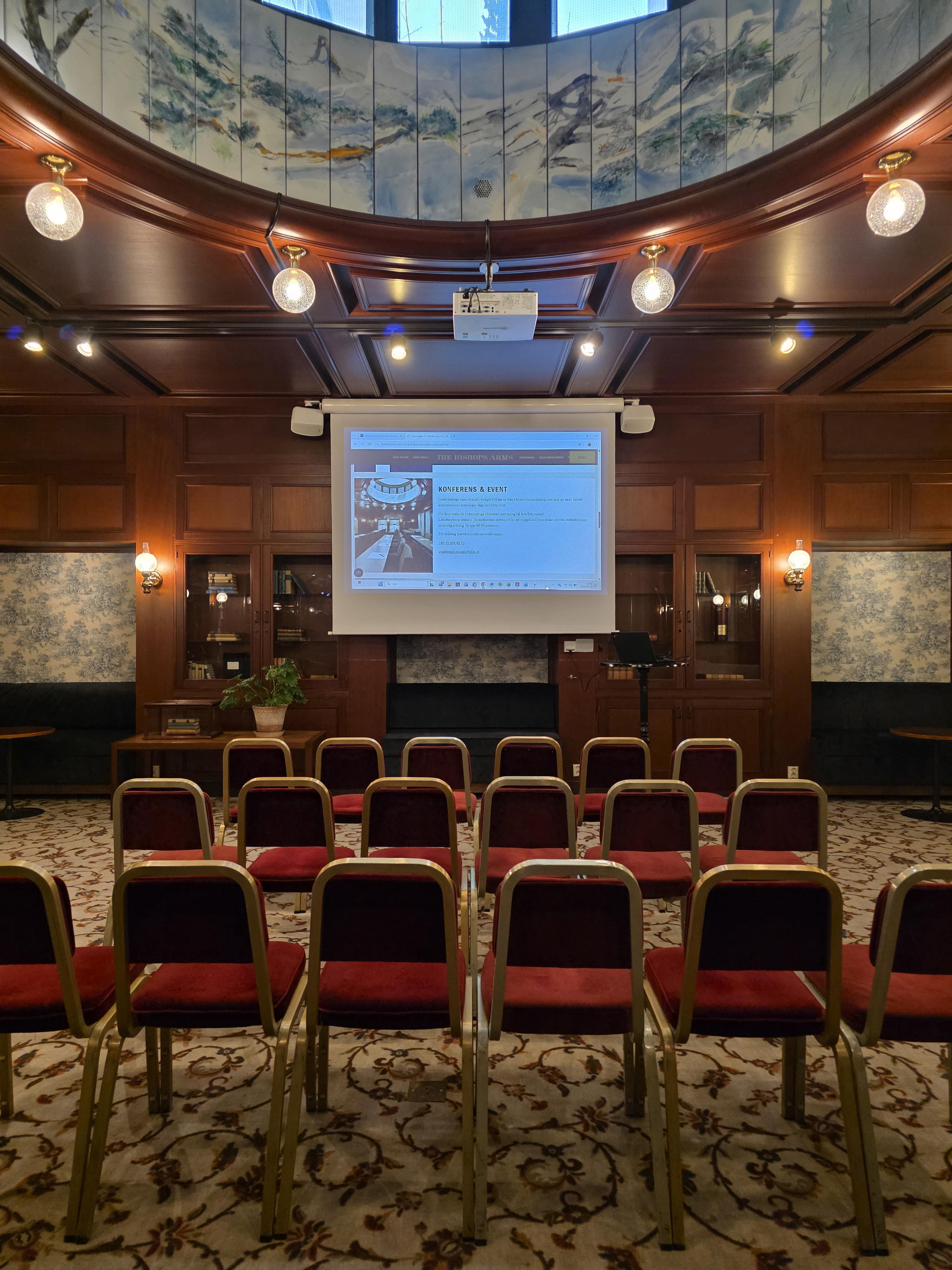 Small conference room with wooden walls, a projector screen displaying a presentation, red cushioned chairs arranged in rows, and decorative lighting above.