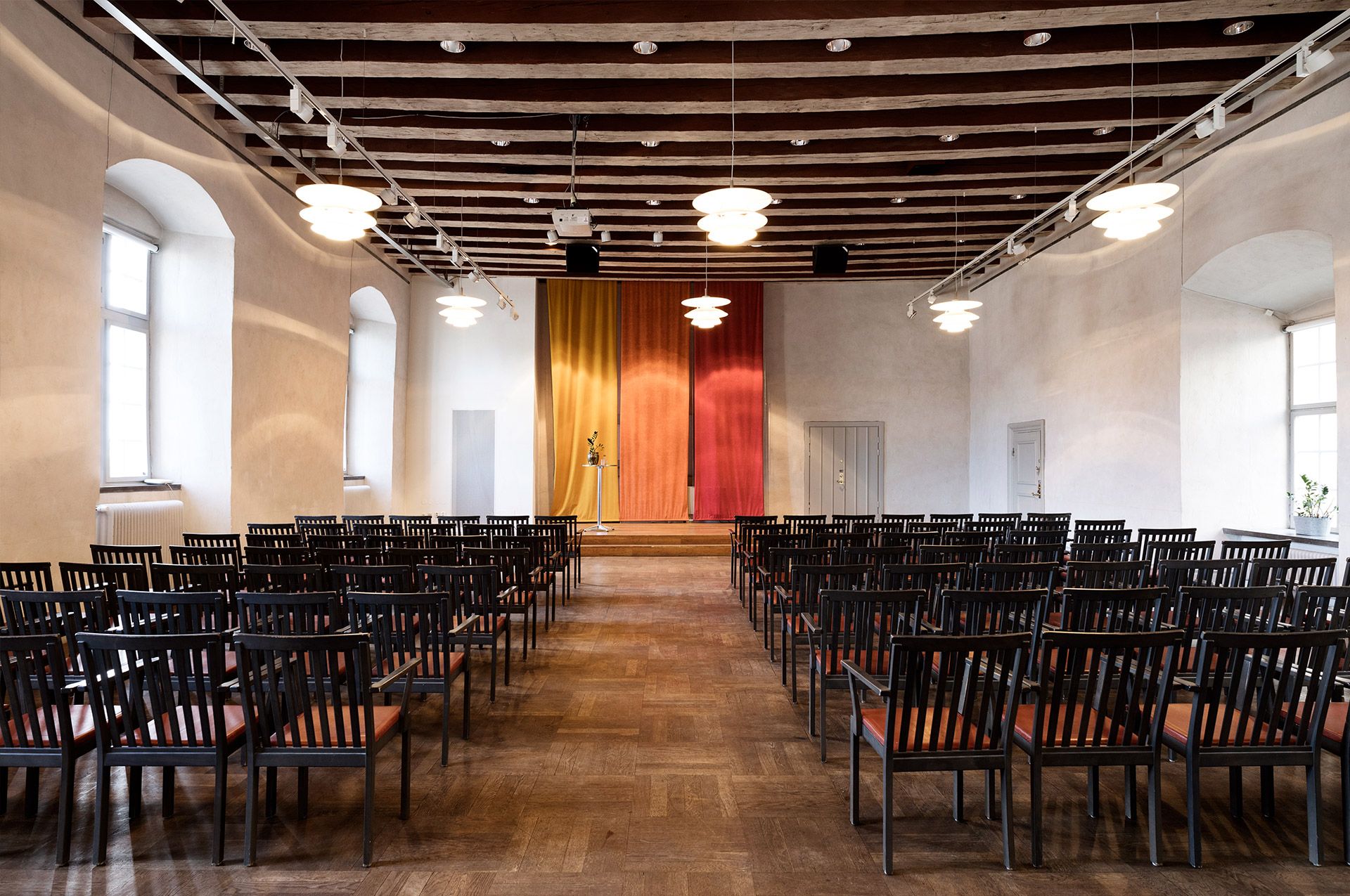 Large room with white walls, windows and lined up black chairs