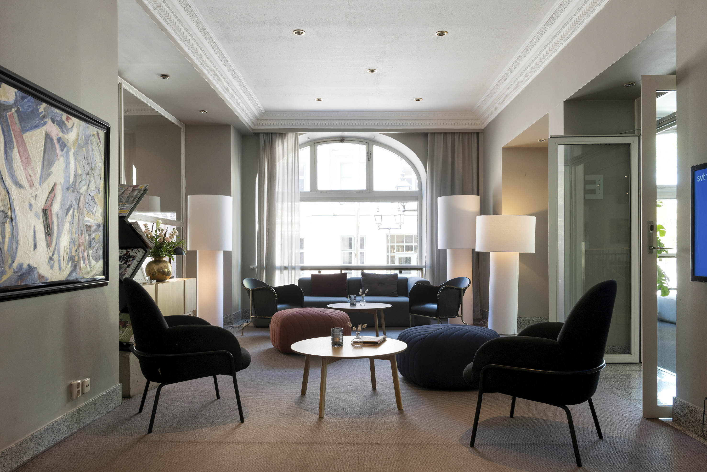 Modern hotel lounge area with stylish black chairs, round coffee tables, large floor lamps, and contemporary artwork. The space is filled with natural light from a large window, creating an inviting atmosphere.