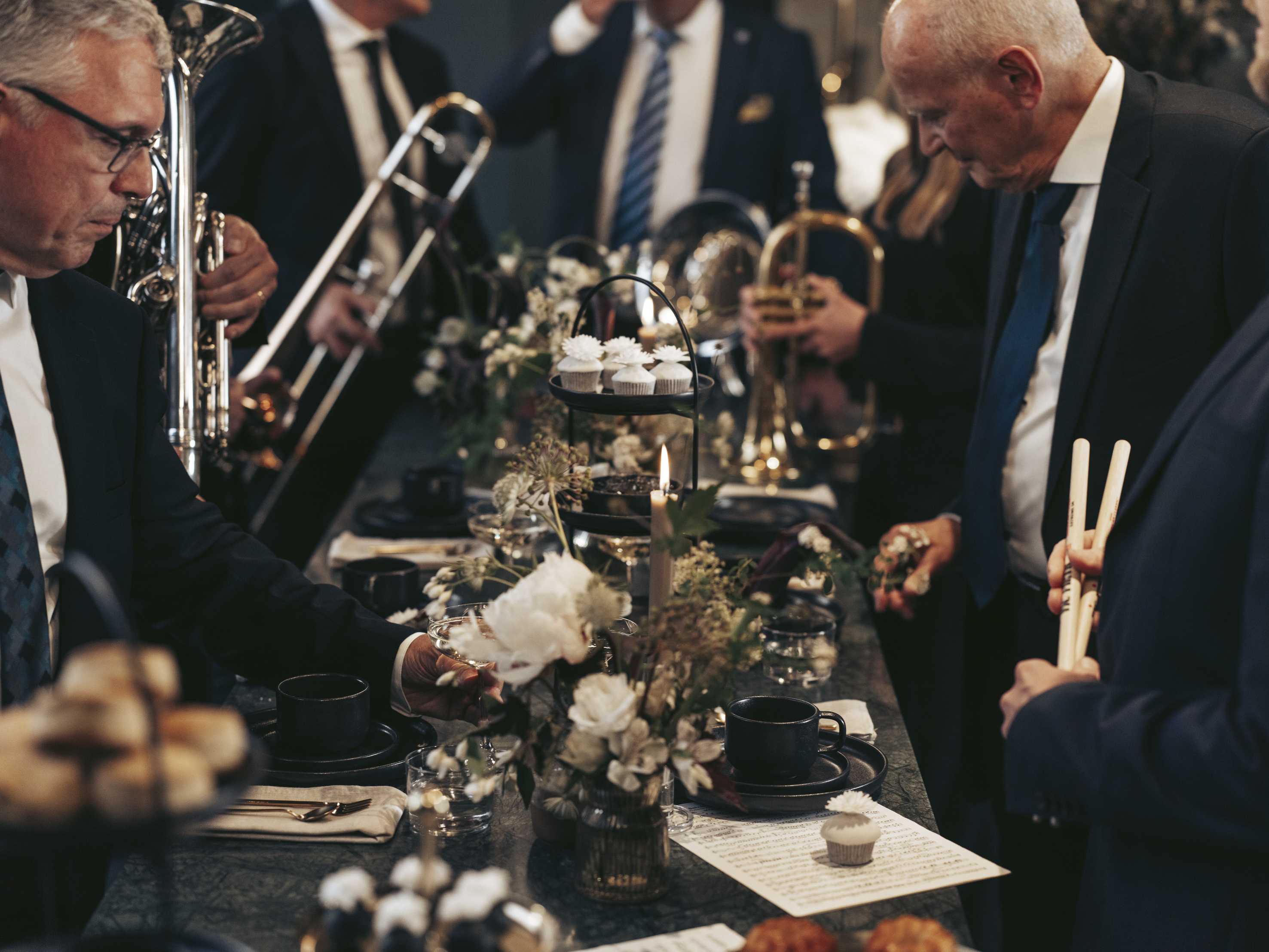 People in formal attire at a festive table setting with flowers and pastries, surrounded by musicians with wind instruments