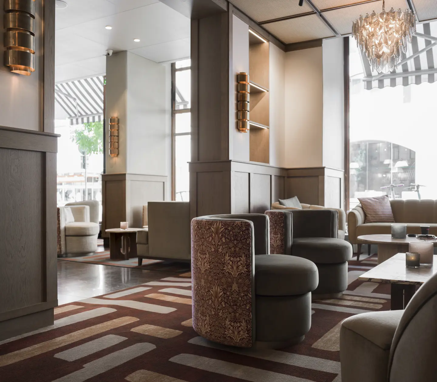 Modern hotel lobby with chic furniture, neutral tones, patterned carpet, and an elegant chandelier, featuring cozy seating areas near large windows and natural light.