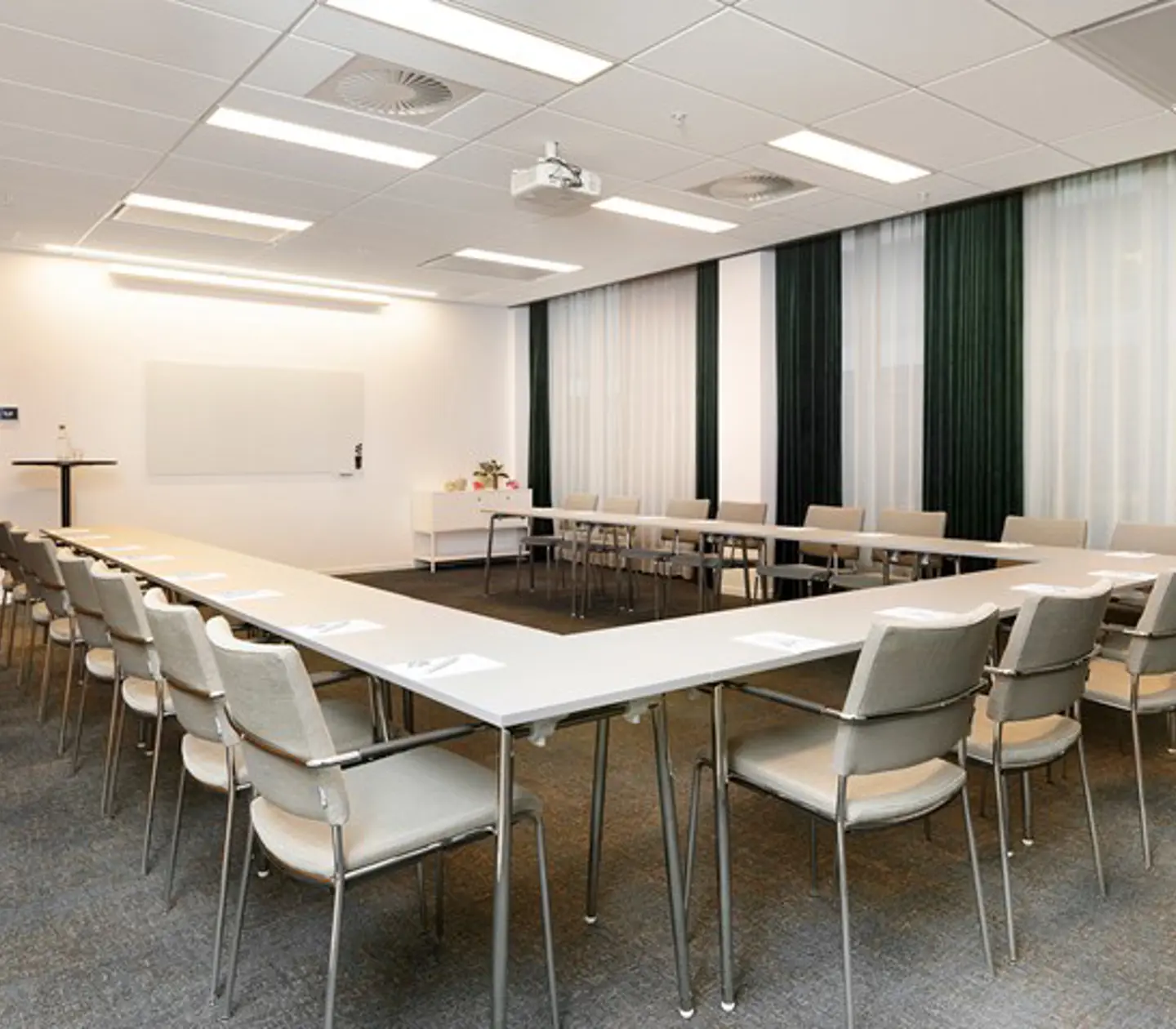 Konferensrum med u-sittning, vita stolar, vitt bord, grå matta, draperier