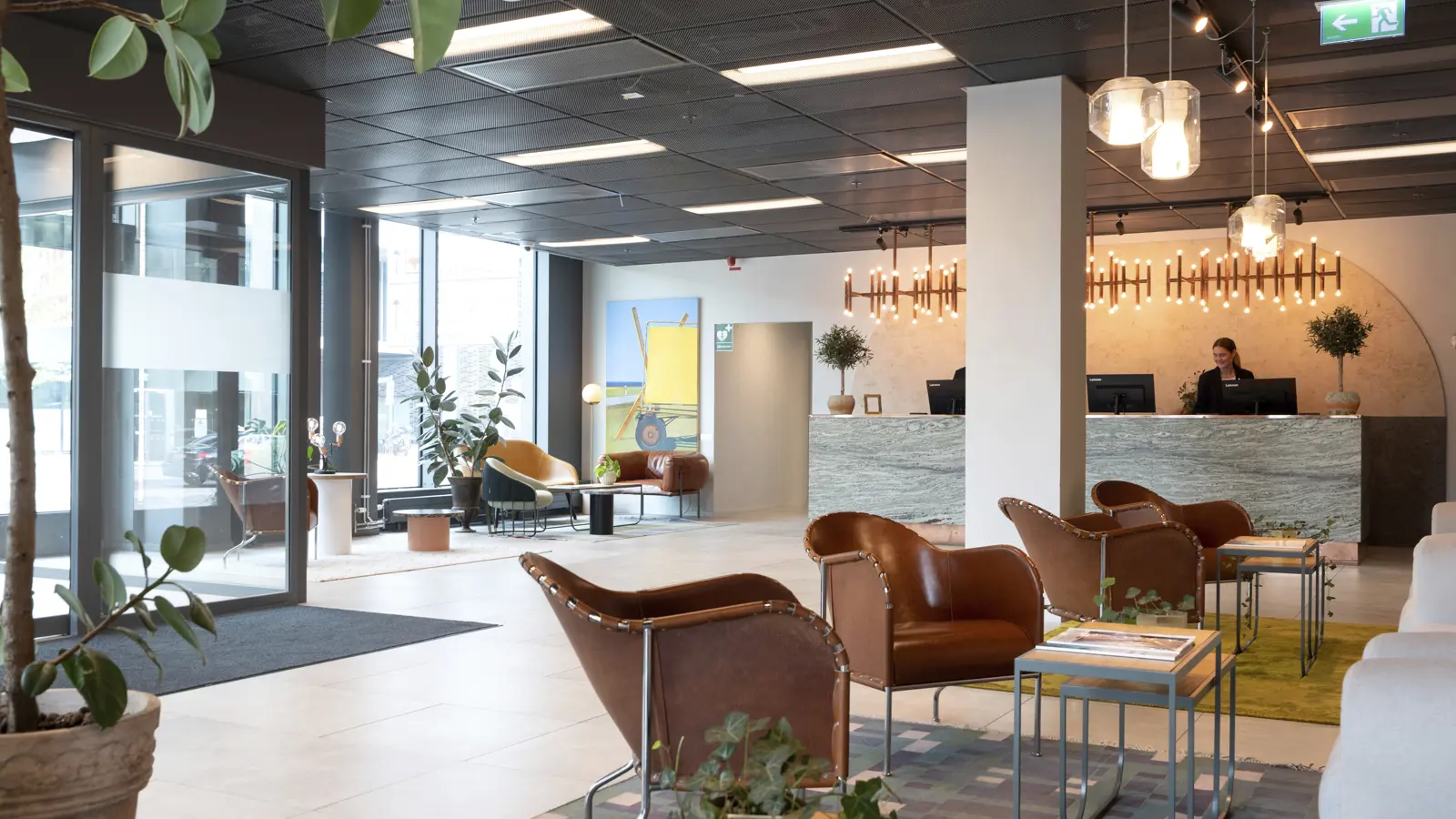 Modern hotellobby med eleganta läderfåtöljer, växter och ljus belysning. Receptionen har en marmordisk och två receptionister. Stora fönster släpper in naturligt ljus och en stilren inredning skapar en inbjudande atmosfär.