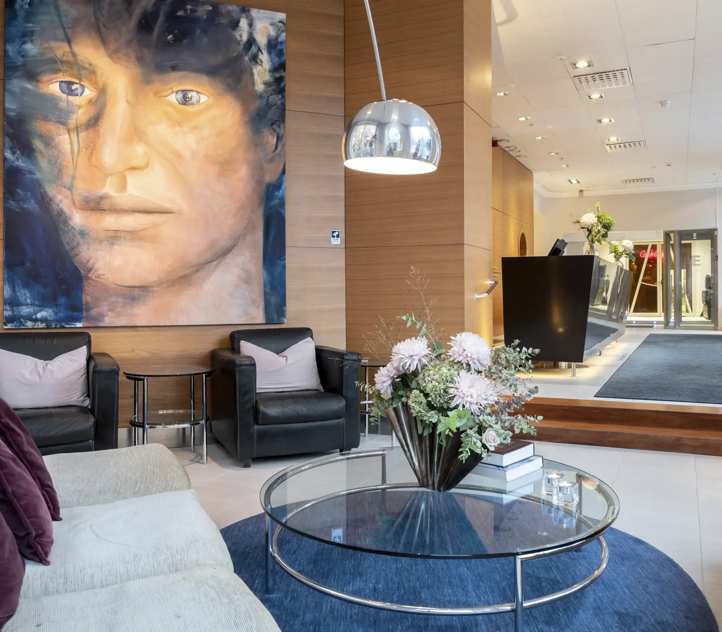 Modern hotel lobby with a large portrait painting on a wooden wall, featuring black leather chairs and a glass coffee table with floral arrangement and books. The reception area has a dark desk and bright overhead lighting.