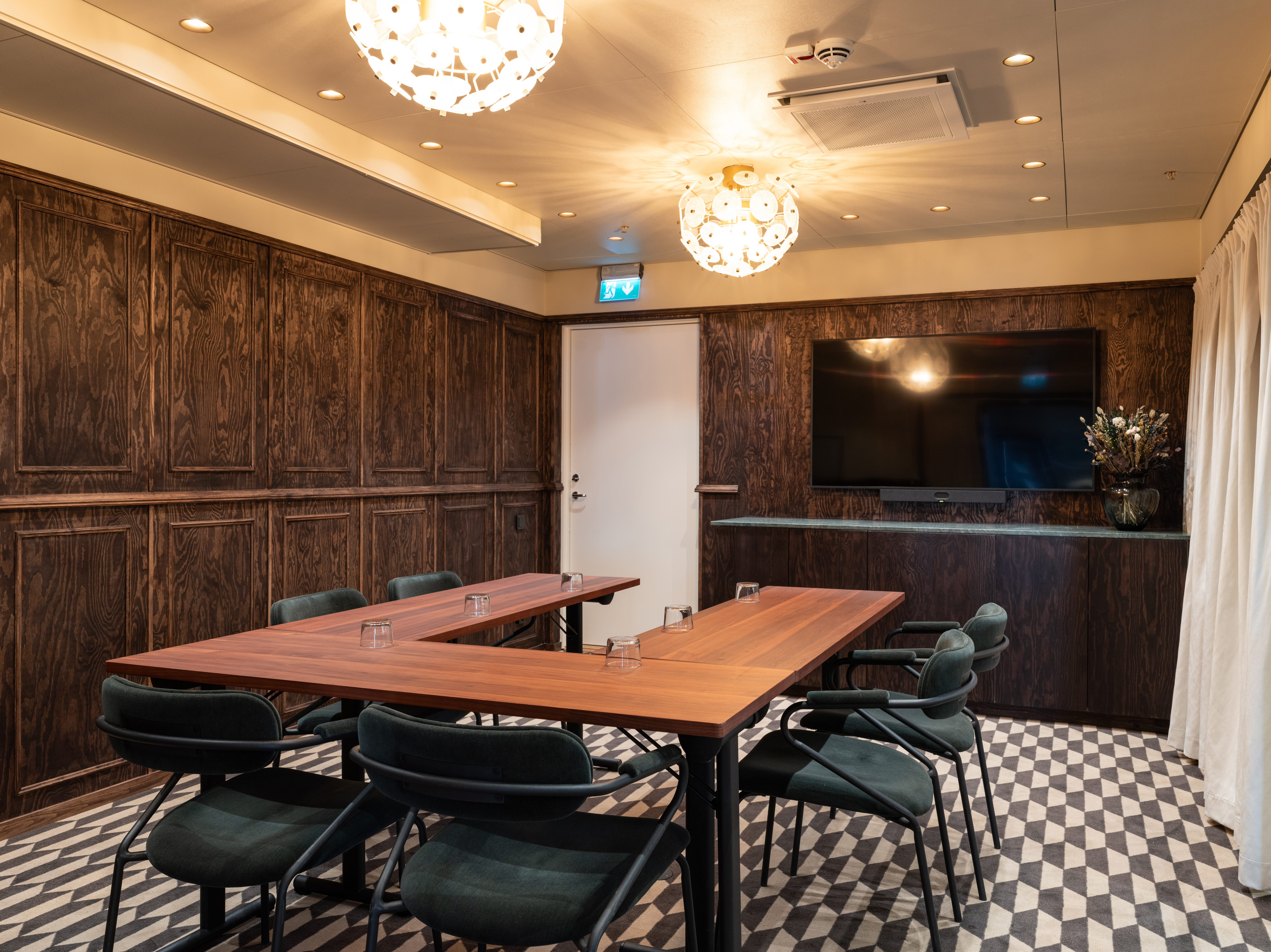 Modern konferensrum med mörkt träpanel, rektangulärt mötesbord, gröna stolar och integrerad TV-skärm.