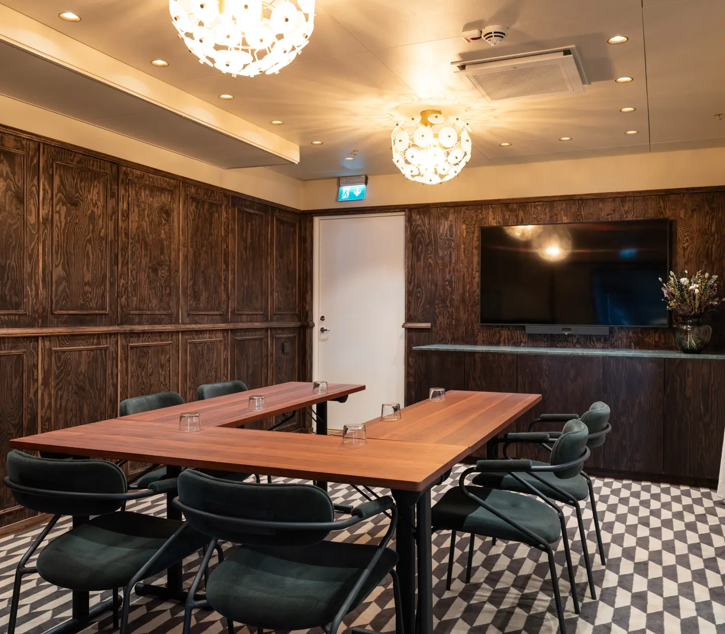 Elegant modern conference room with wooden panel walls, a large rectangular table, green upholstered chairs, geometric patterned floor, and decorative lighting fixtures.
