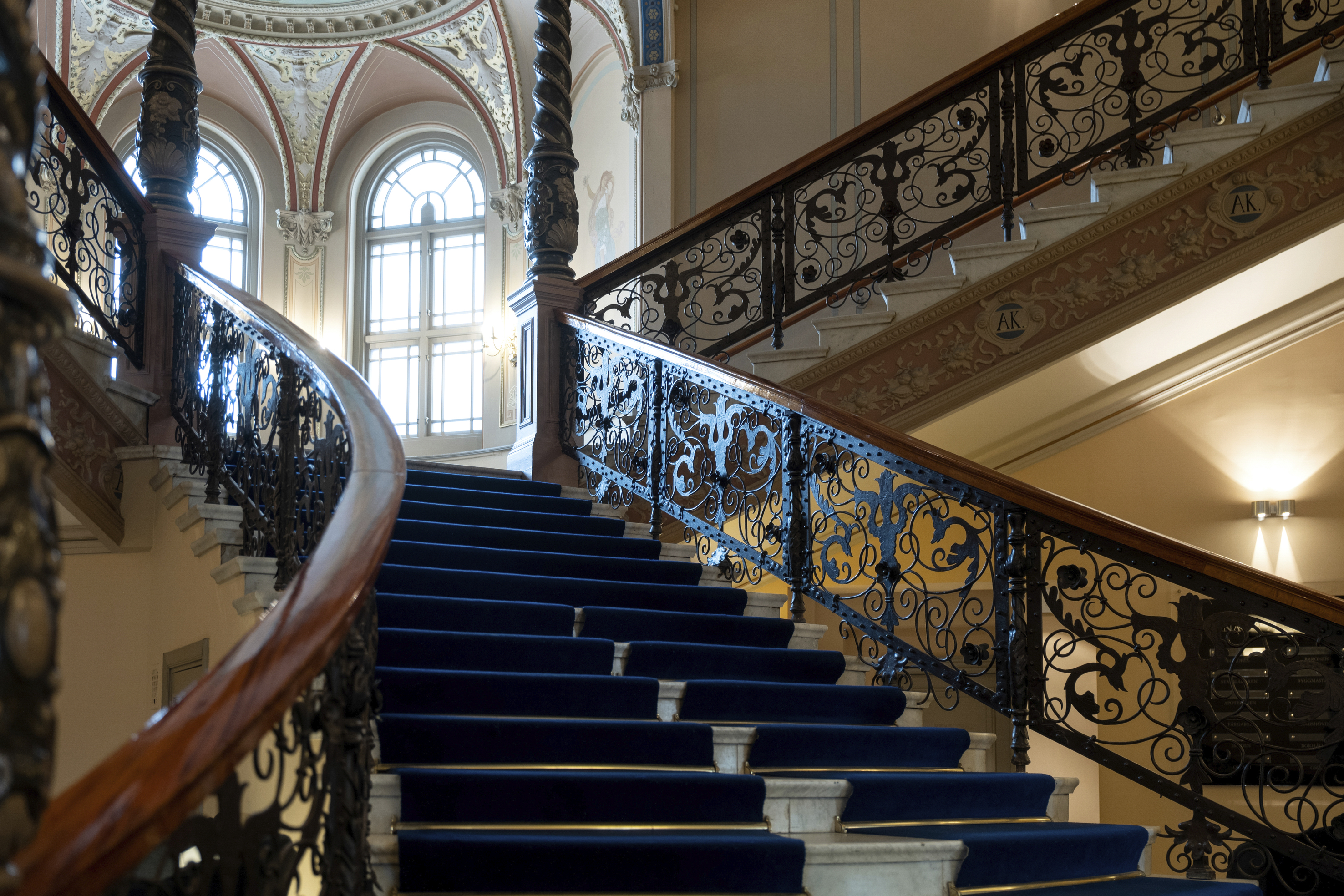 Ornate staircase with blue carpet, intricate wrought iron railing, and decorative arched windows in an elegant interior setting.