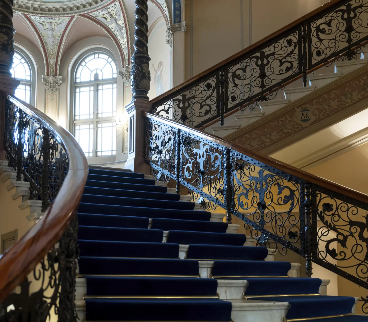 Ornate staircase with blue carpet, intricate wrought iron railing, and decorative arched windows in an elegant interior setting.