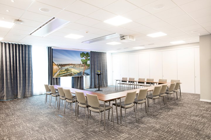 Konferensrum med u-sittning, grått golv, grå stolar och tända lampor i taket