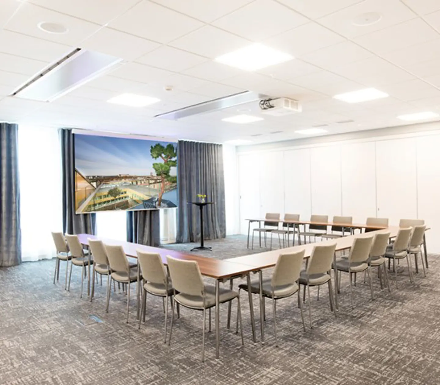 Konferensrum med u-sittning, grått golv, grå stolar och tända lampor i taket
