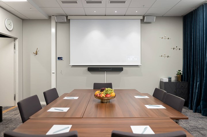 Konferensrum med brunt bord, grå stolar, ljusa väggar och projektor