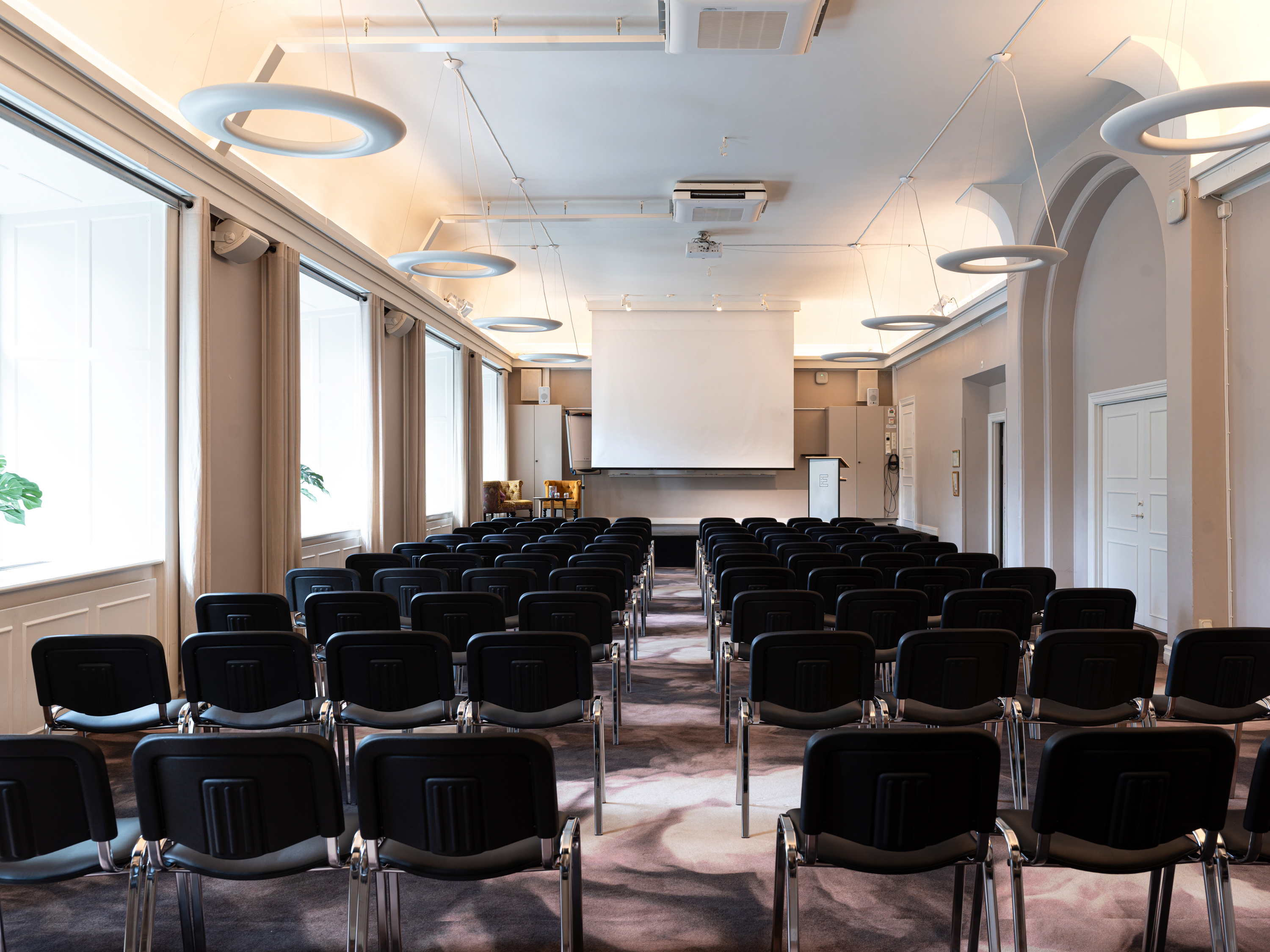 Konferensrum med rader av svarta stolar, projektorduk och modern belysning. Perfekt för möten och presentationer.