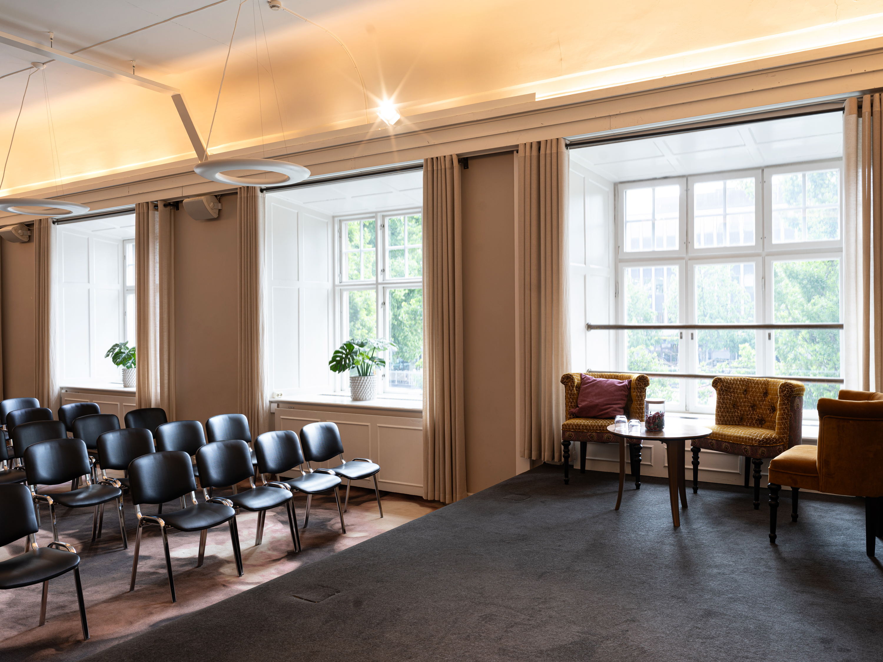 Konferensrum med stolar och stora fönster, växter och två fåtöljer med ett litet bord, i ett modernt mötesutrymme.