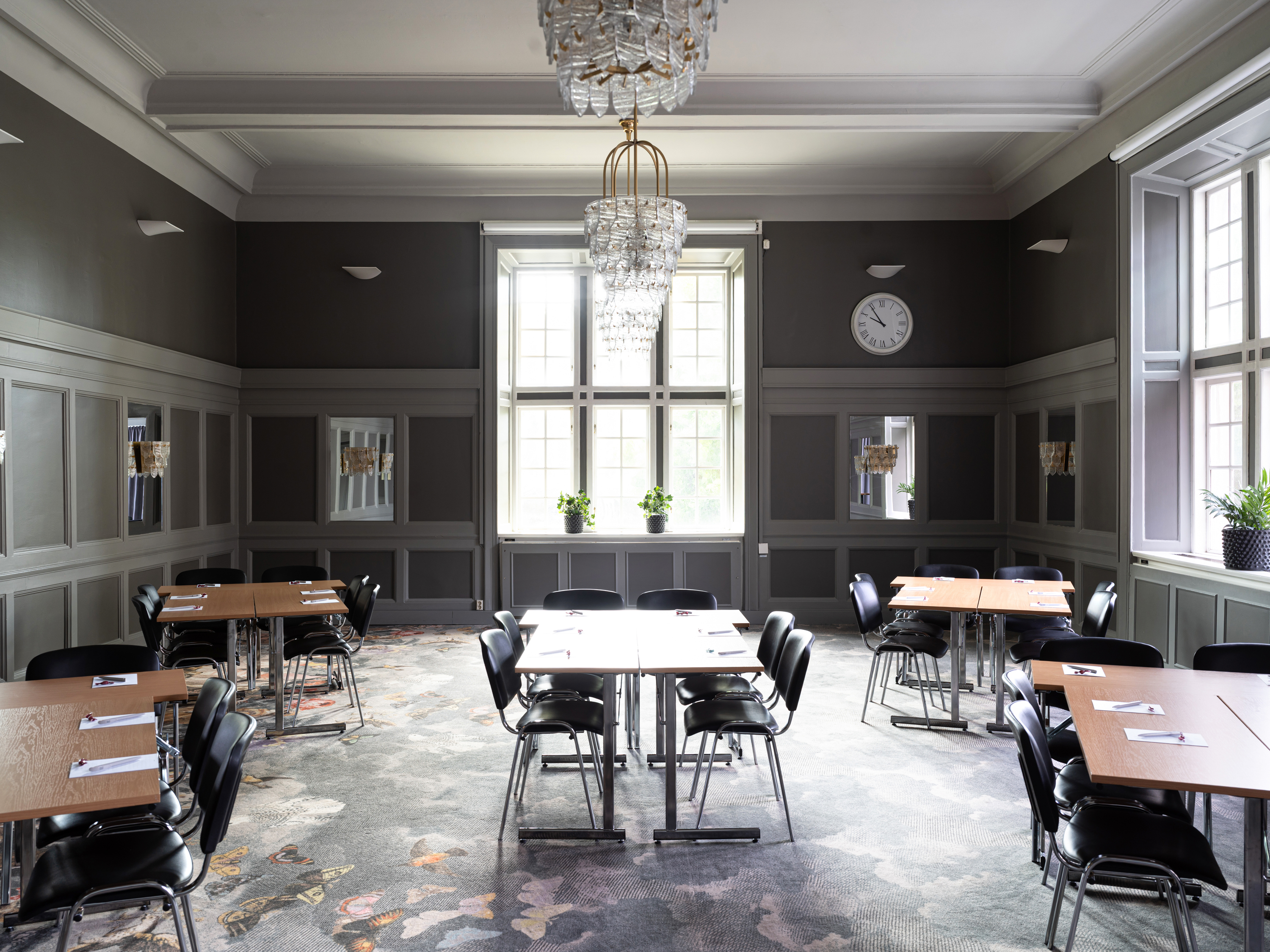 Ljust och elegant mötesrum med moderna möbler, stort fönster med naturligt ljus, väggklocka och dekorativa växter.