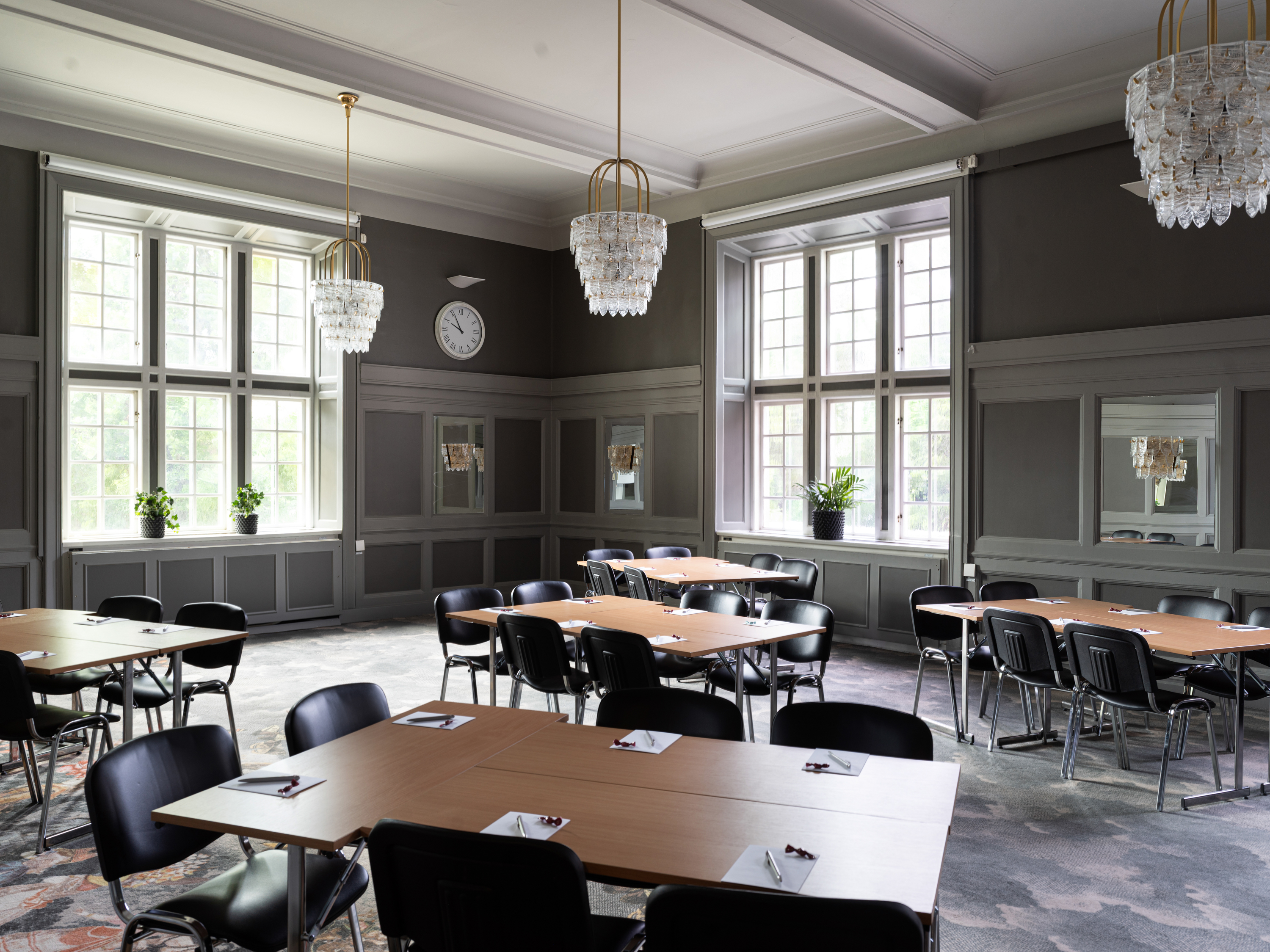 Konferensrum med grå väggar, stora fönster och eleganta ljuskronor, möblerat med bord och stolar, perfekt för möten och arbete.