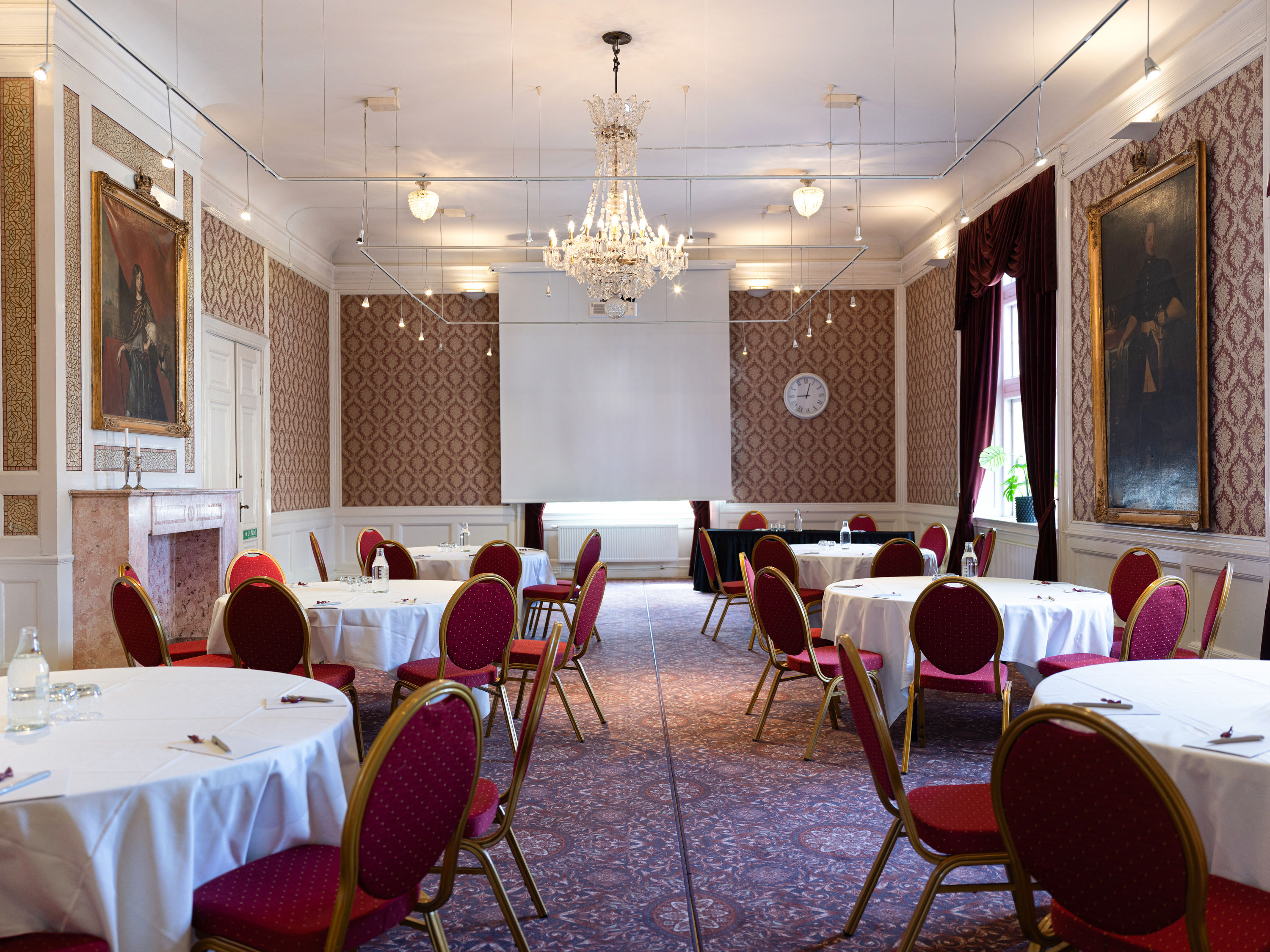 Konferenssal med runda bord och röda stolar, elegant inredning med kristallkrona och konstverk på väggarna.