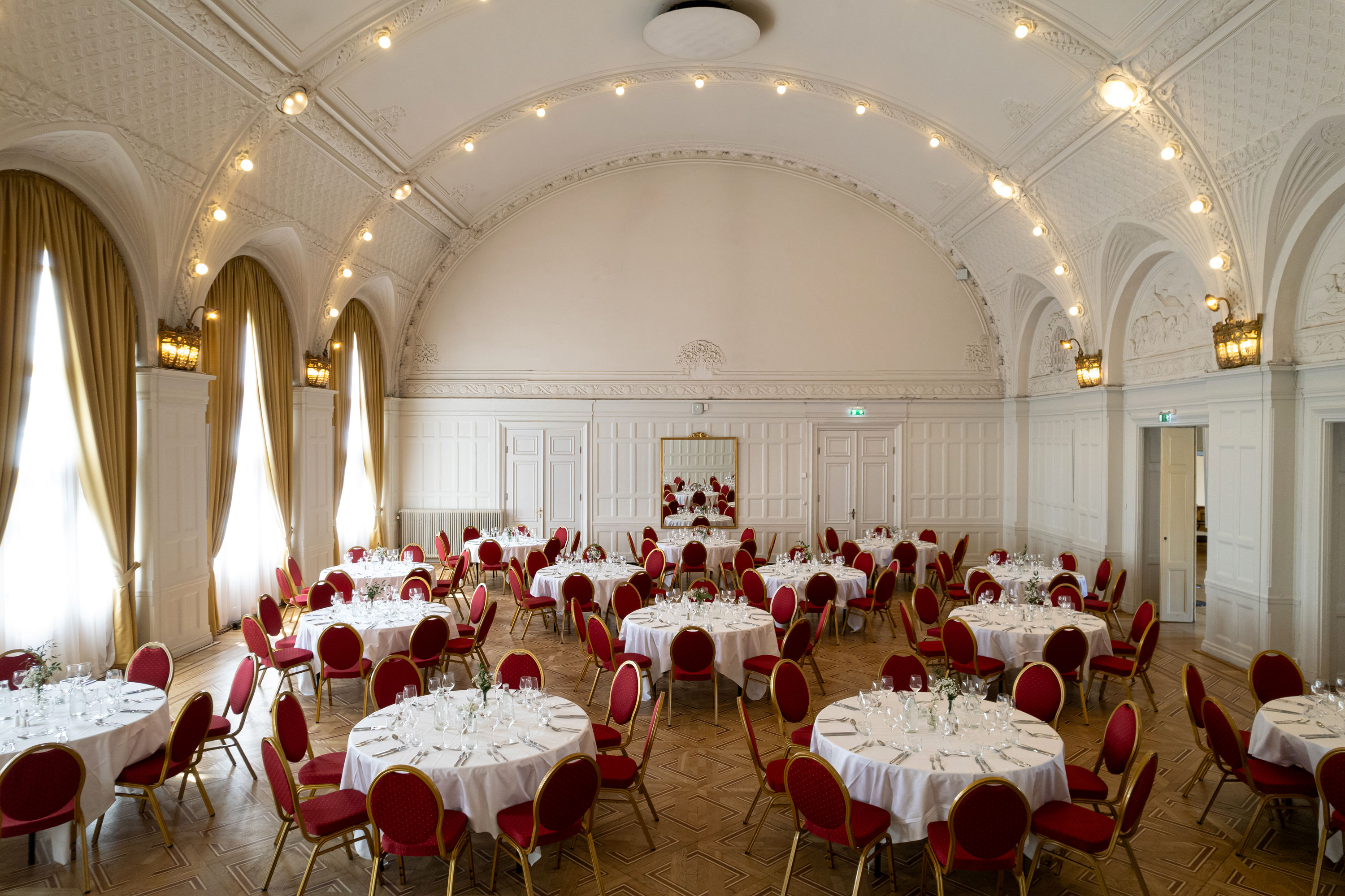 Elegant festsal med vita väggar och högt i tak, dekorerad med runda bankettbord, röda stolar och dukar.