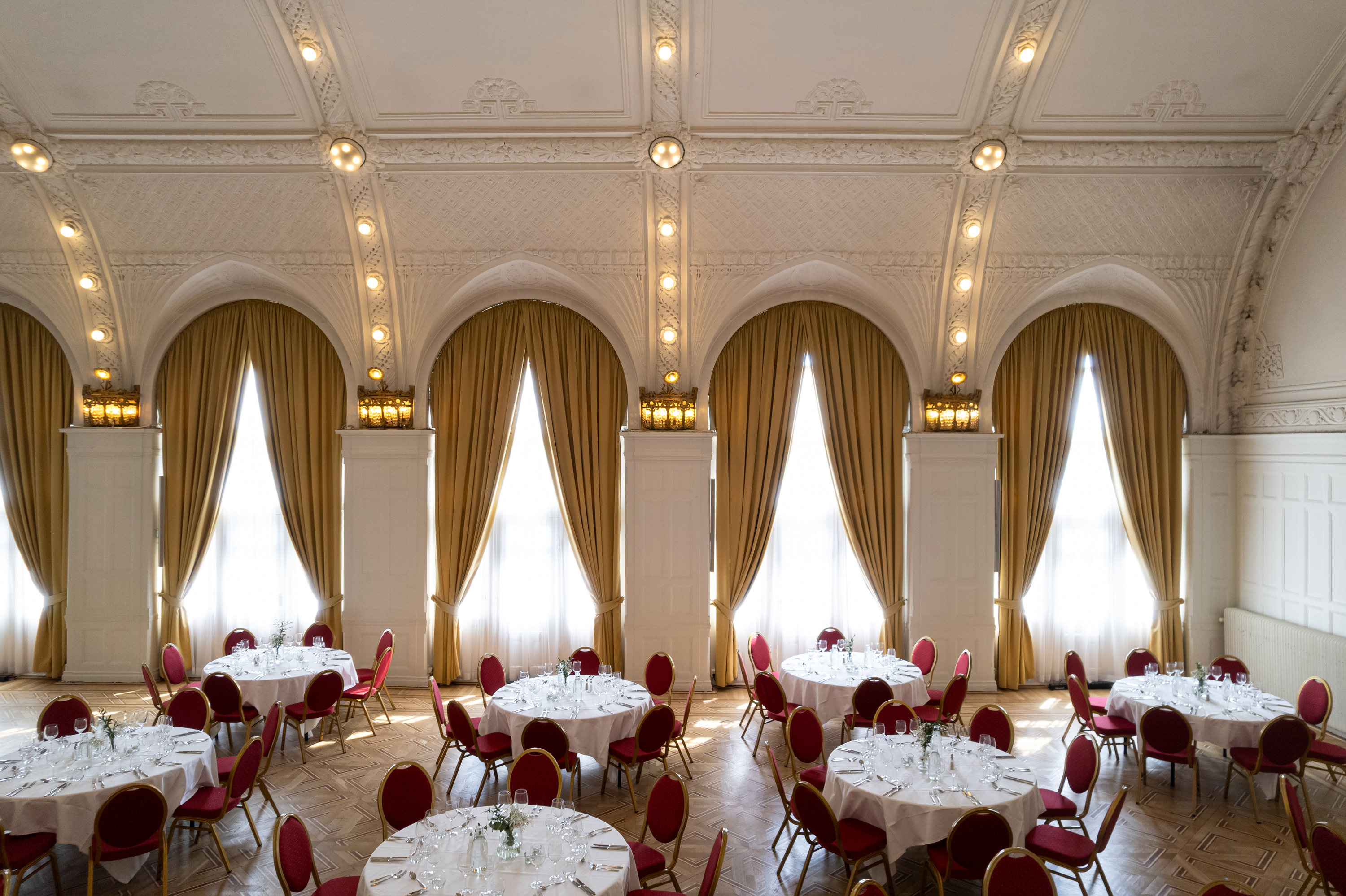 Elegant bankettsal med rundade bord, röda stolar och höga fönster med gyllene draperier. Historisk arkitektur med detaljerat innertak och mjuk belysning.