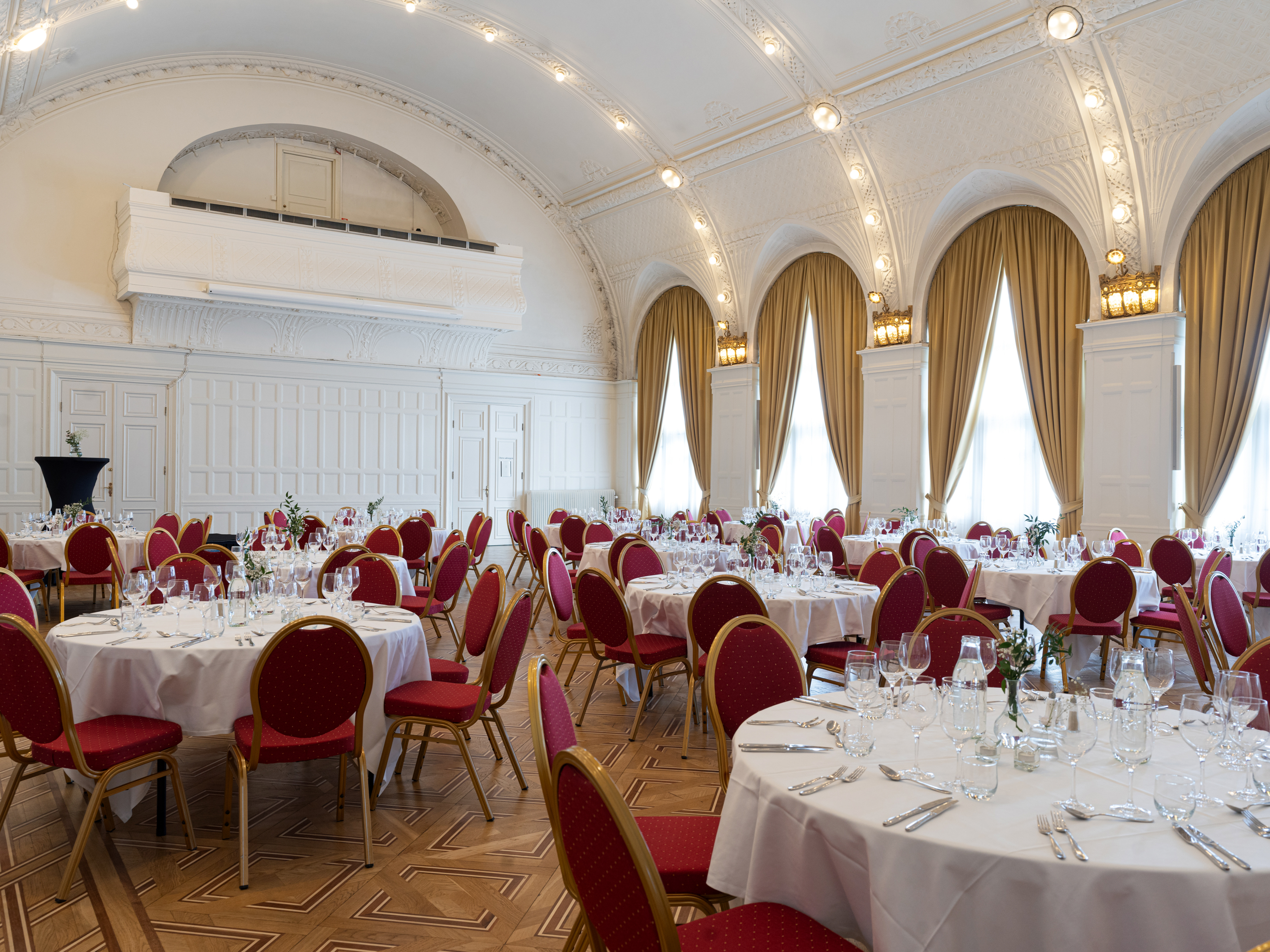 Elegant bankettsal med uppdukat för fest, vita dukar och röda stolar, stora fönster med gyllene gardiner och dekorativa lampor.