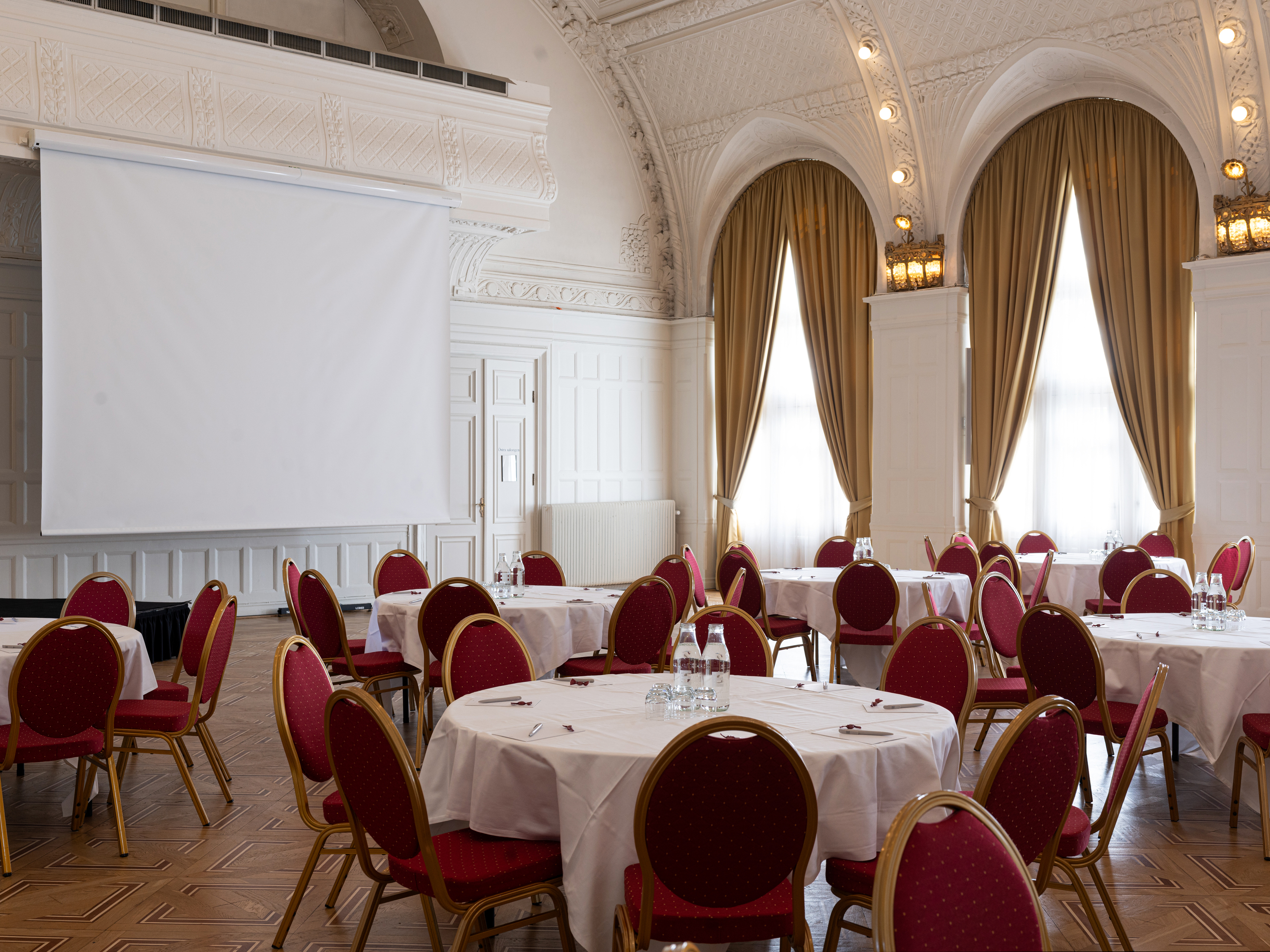 Elegant mötesrum med välvda tak, runda bord dukade med vita dukar och röda stolar, projektorduk och högre fönster med gardiner.