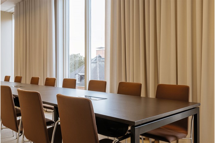 Konferensrum med styrelsesittning, brunt bord, bruna stolar, stora fönster och ljusa draperier