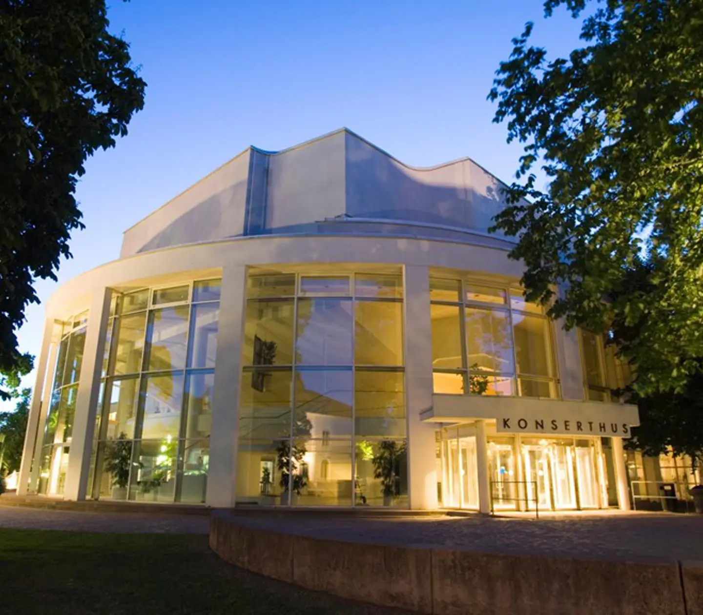 Modern concert hall exterior illuminated at dusk, surrounded by trees, with large glass windows and "Konserthus" sign at entrance.