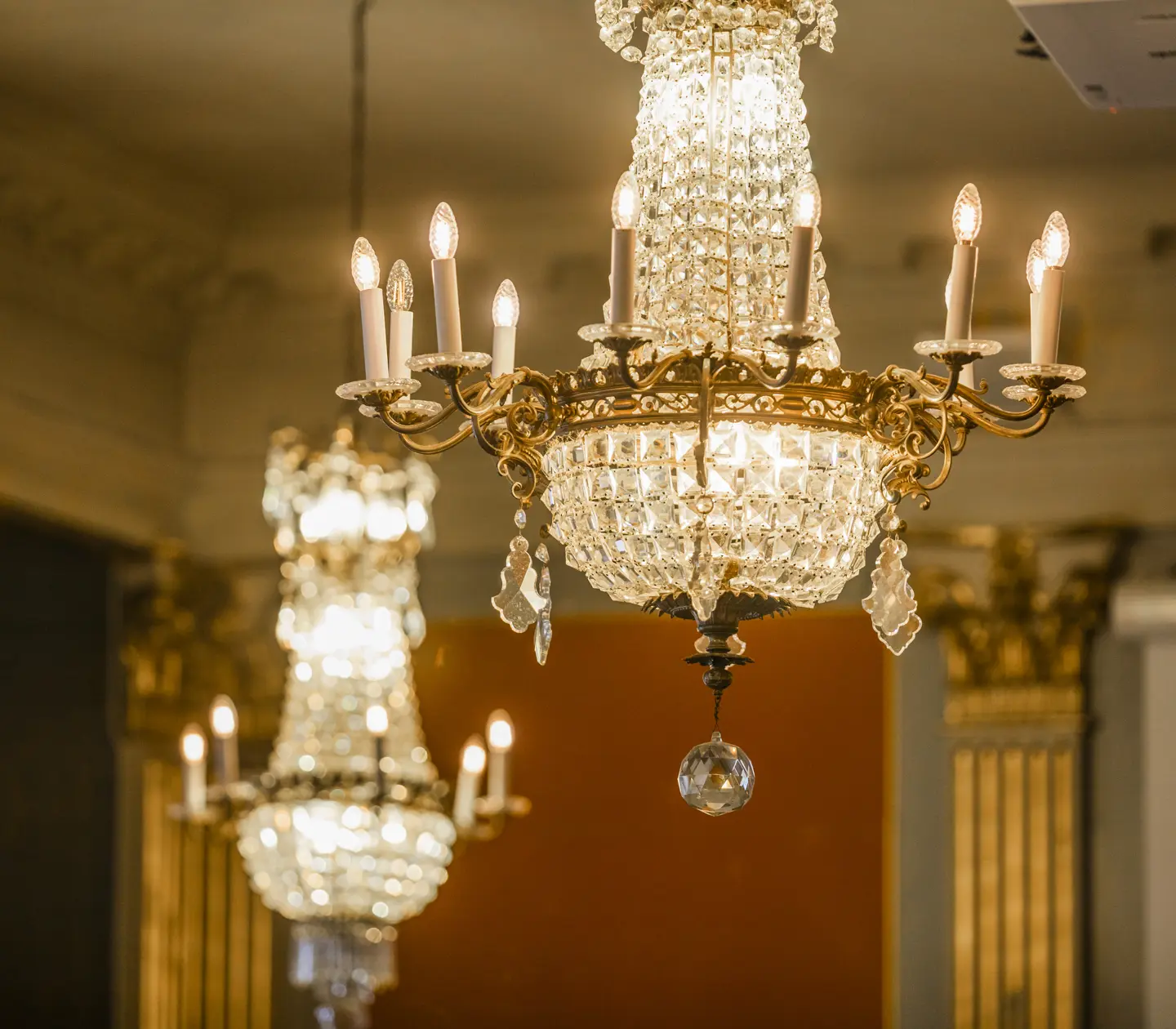 Elegant crystal chandeliers with lit candles in a grand ballroom setting, featuring ornate gold detailing and ambient lighting.