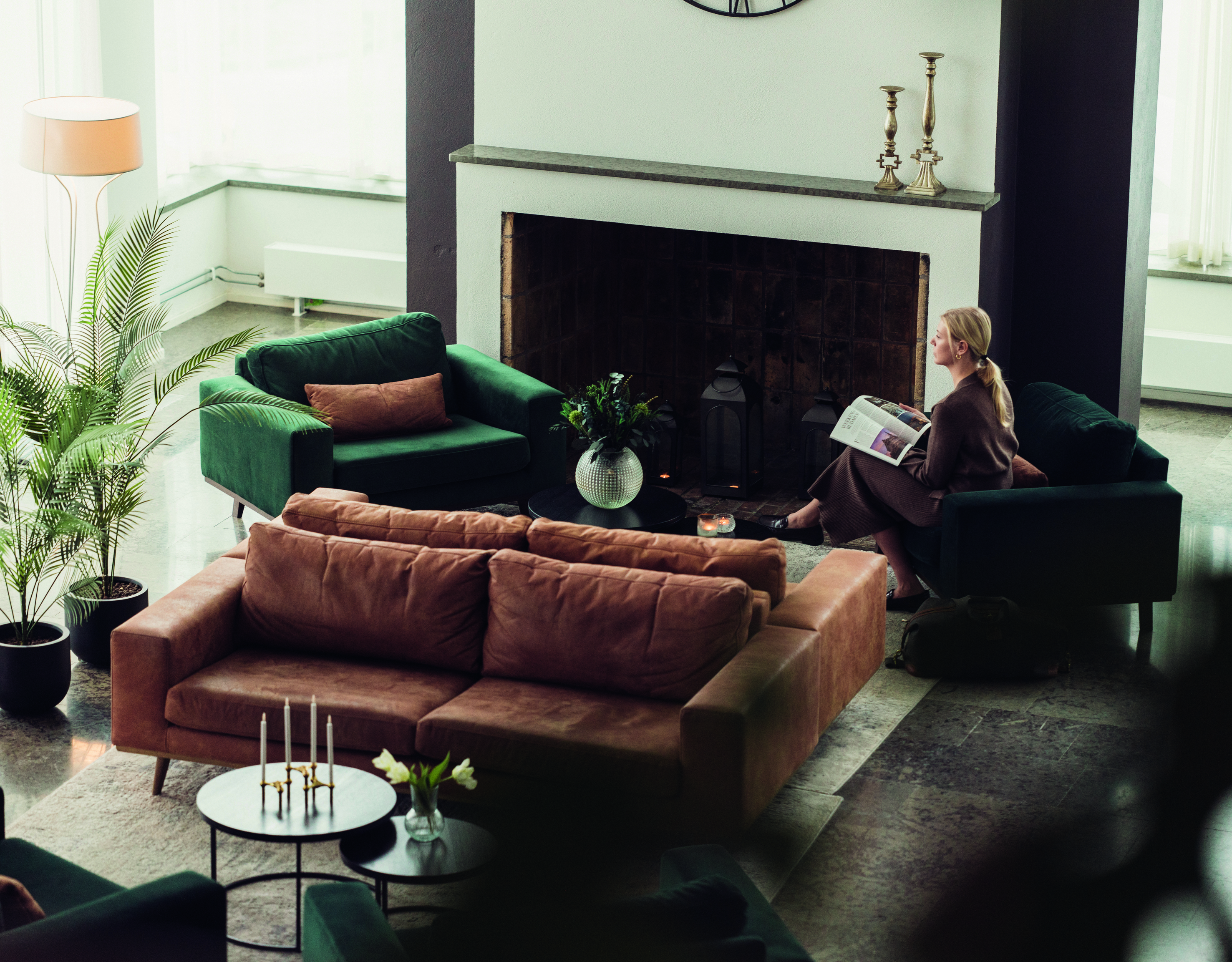Modern living room with green and brown sofas, a fireplace, indoor plants, and a person reading by the fire.