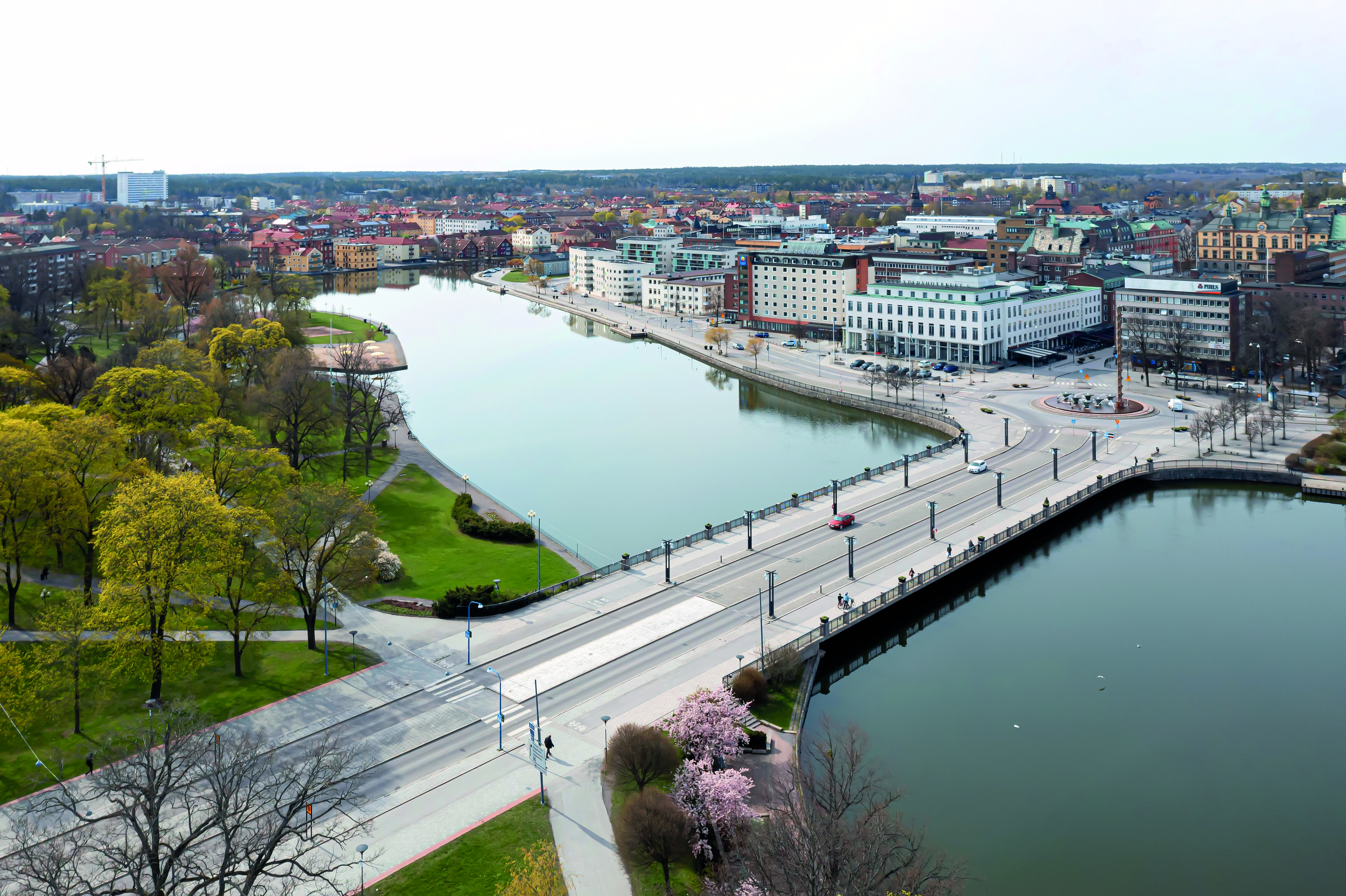 Aerial view of a cityscape featuring a calm river flanked by green parks and urban buildings, with a prominent bridge crossing the river.