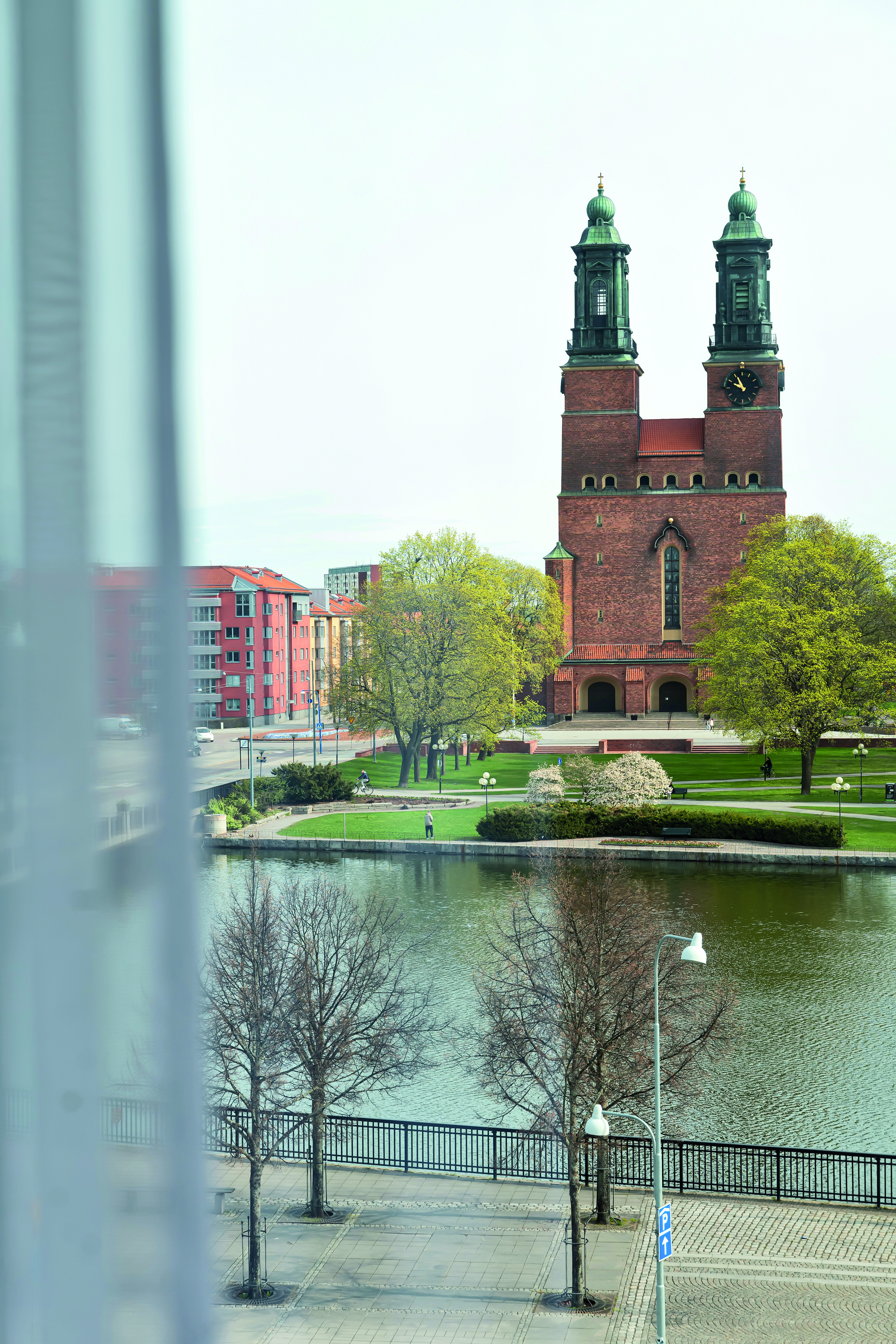 View of a historic brick church with twin towers by a river, surrounded by green trees and lawns, in a cityscape with modern buildings in the background.