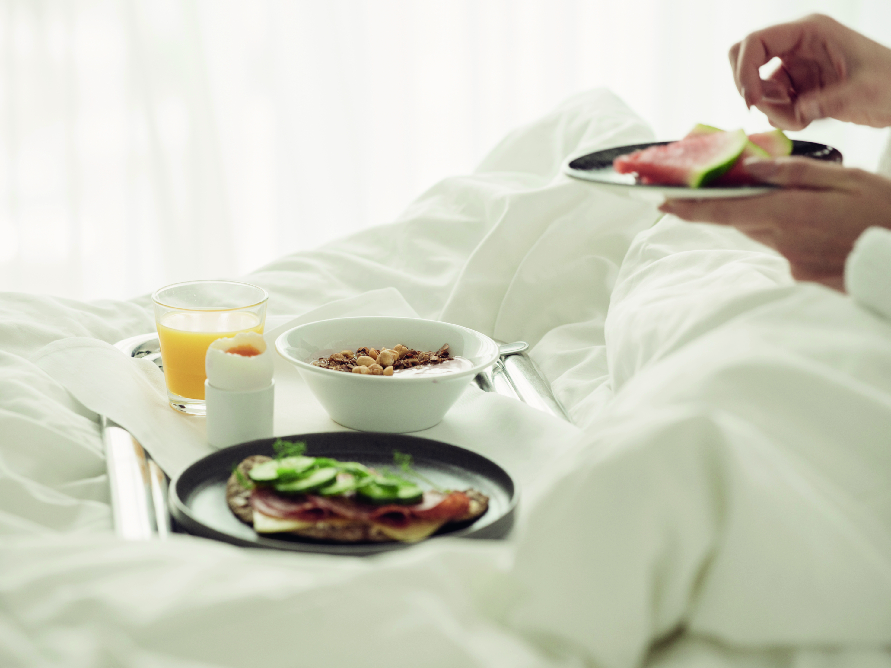 Breakfast in bed with a tray featuring orange juice, yogurt with granola, a sandwich with cucumbers, and a person holding a plate of watermelon slices.
