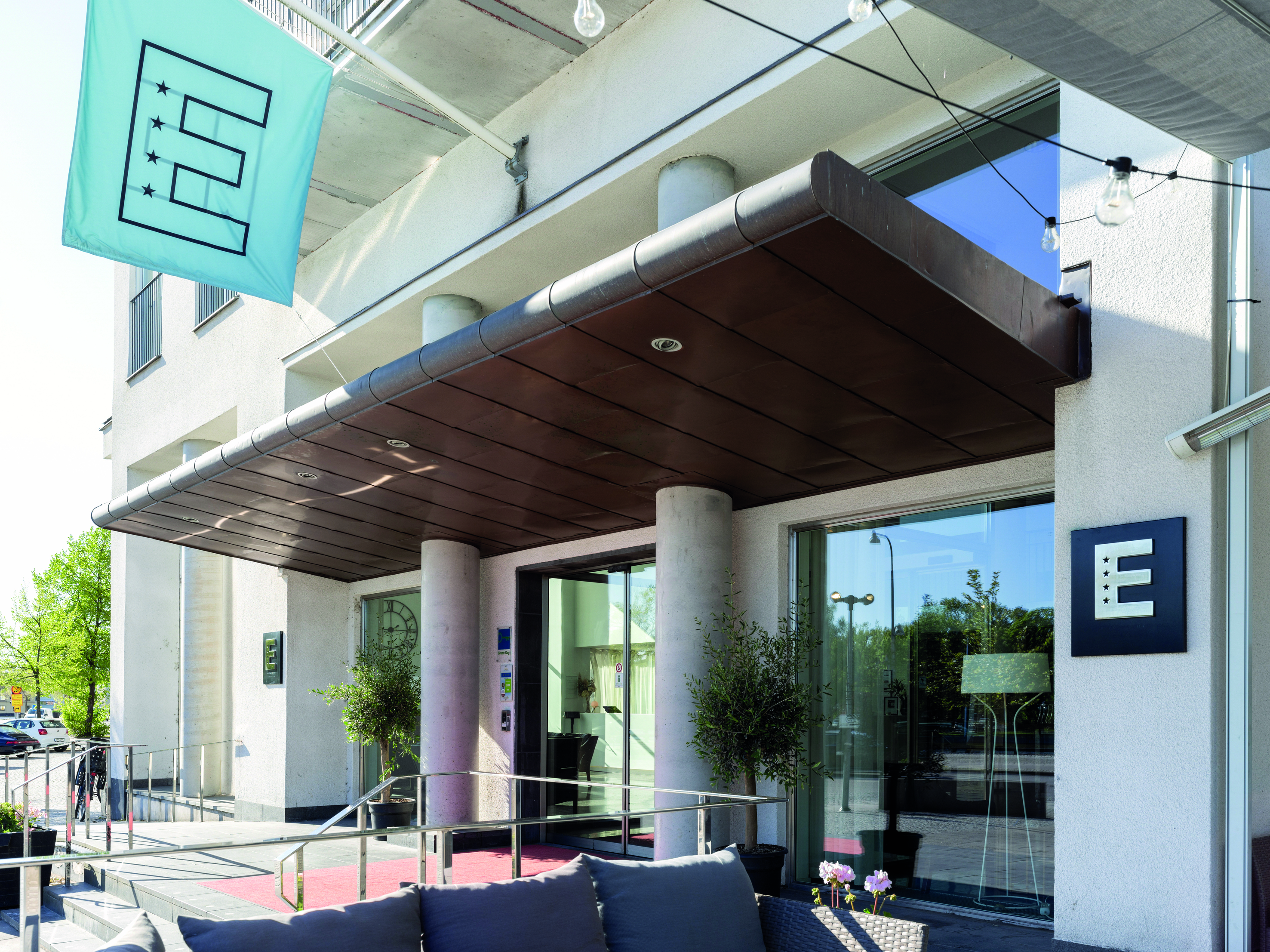 Modern hotel entrance with a teal flag displaying the letter "E", featuring a covered walkway and large glass doors.