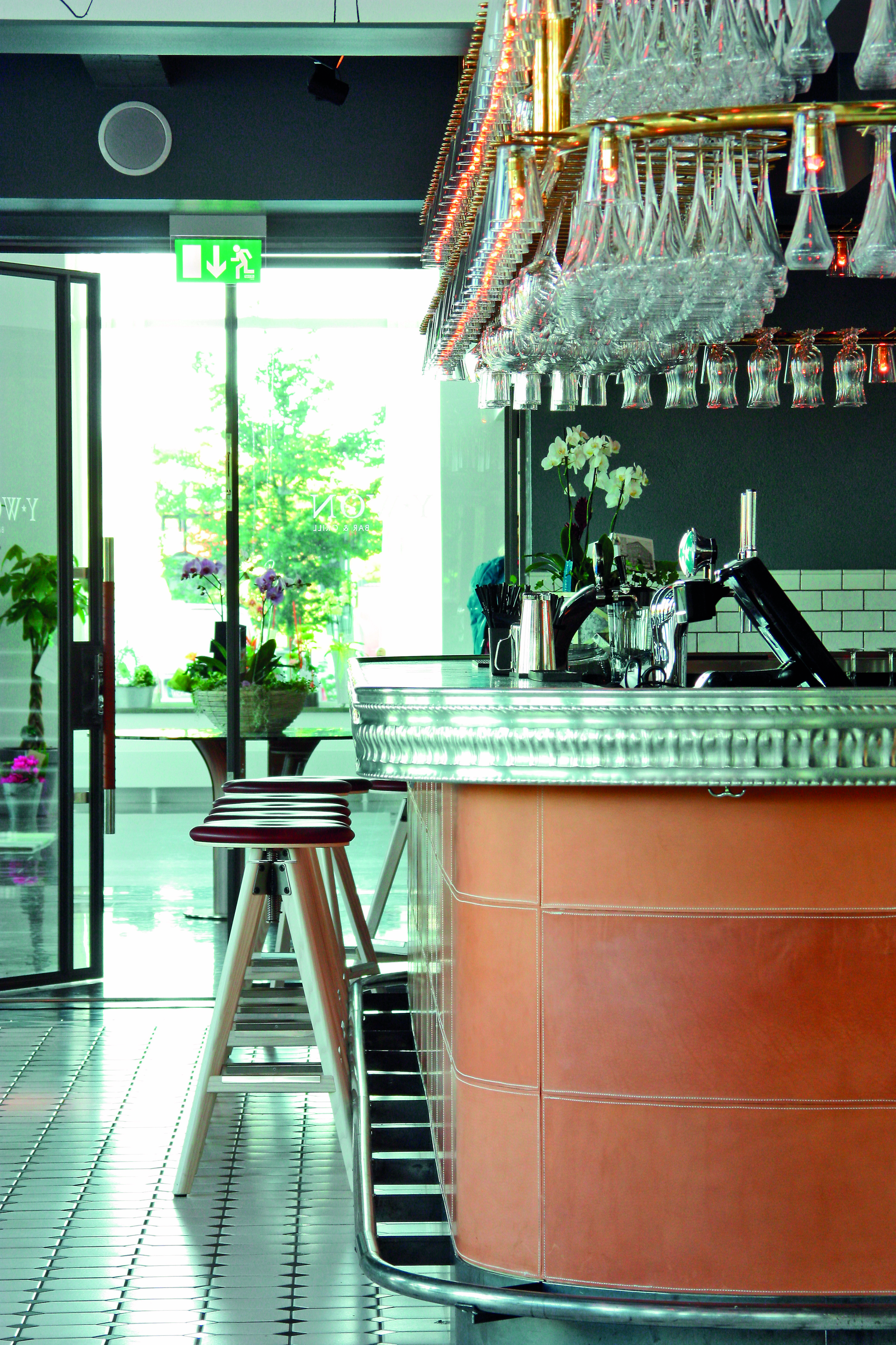 Modern bar interior with sleek counter, hanging wine glasses, and wooden stools, featuring bright, open doorway and greenery.