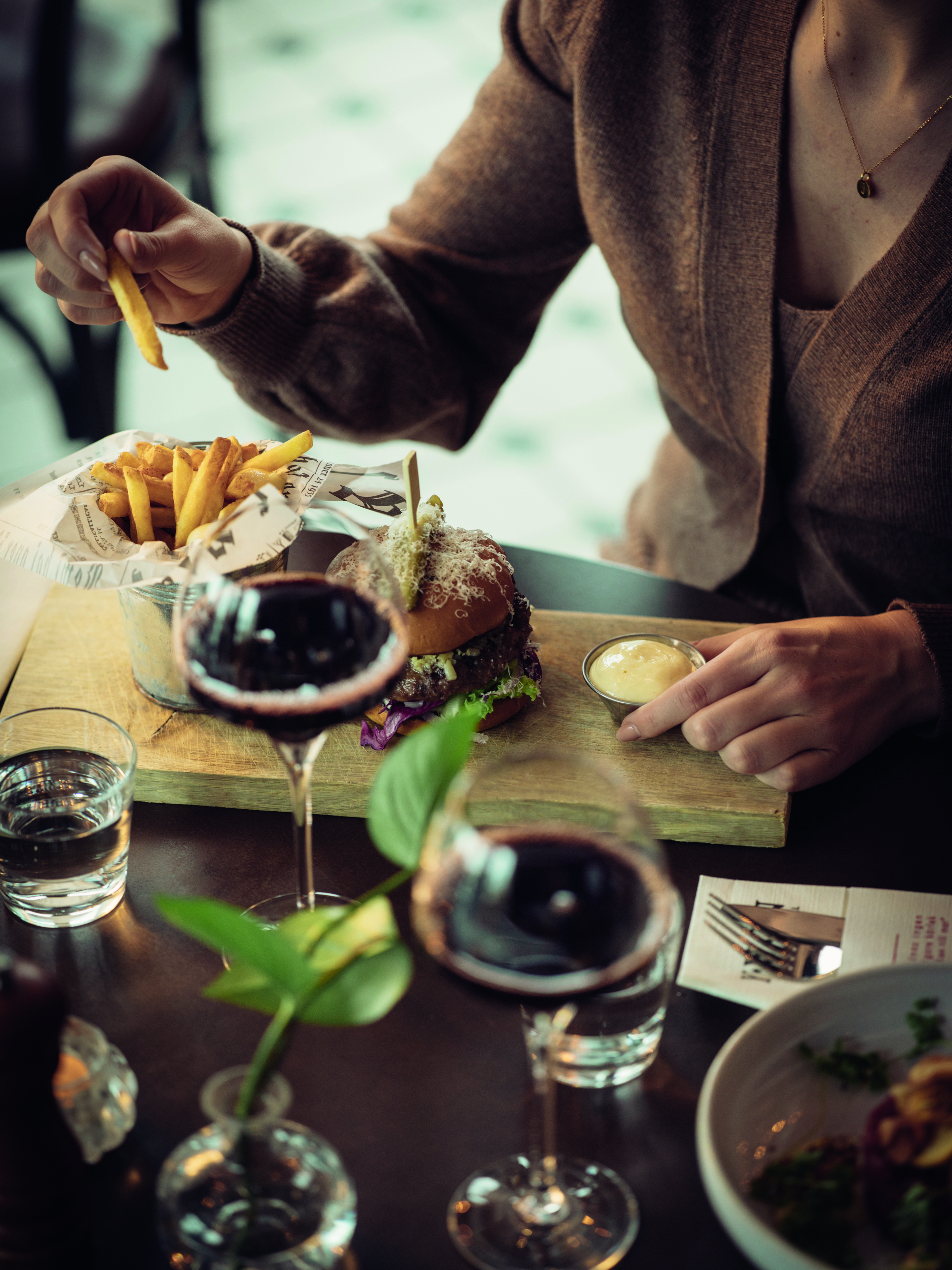 Person enjoying a gourmet burger and fries with red wine at a restaurant table, featuring a wooden serving board and elegant glassware.