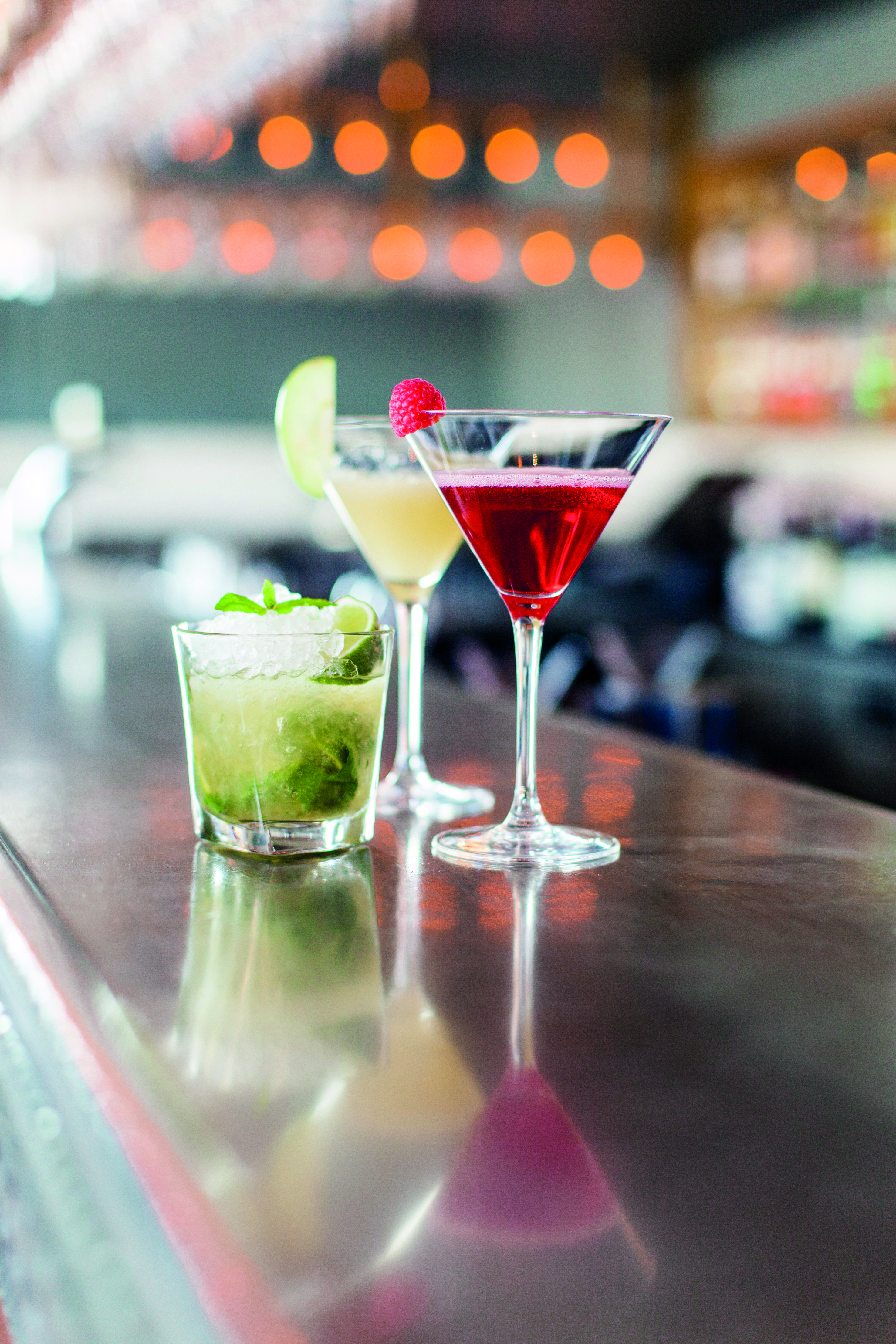 Three stylish cocktails on a bar counter, featuring a mojito garnished with mint and lime, a yellow cocktail with a lime slice, and a red cocktail with a raspberry, set against a blurred bar background.