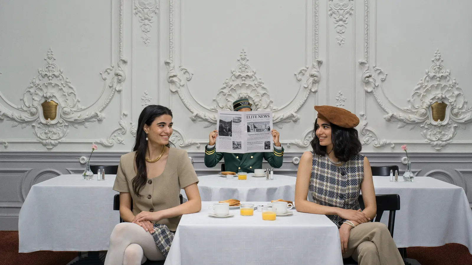 Två kvinnor sitter vid ett elegant dukat bord i en klassisk, dekorativ matsal medan en person läser en tidning i bakgrunden.