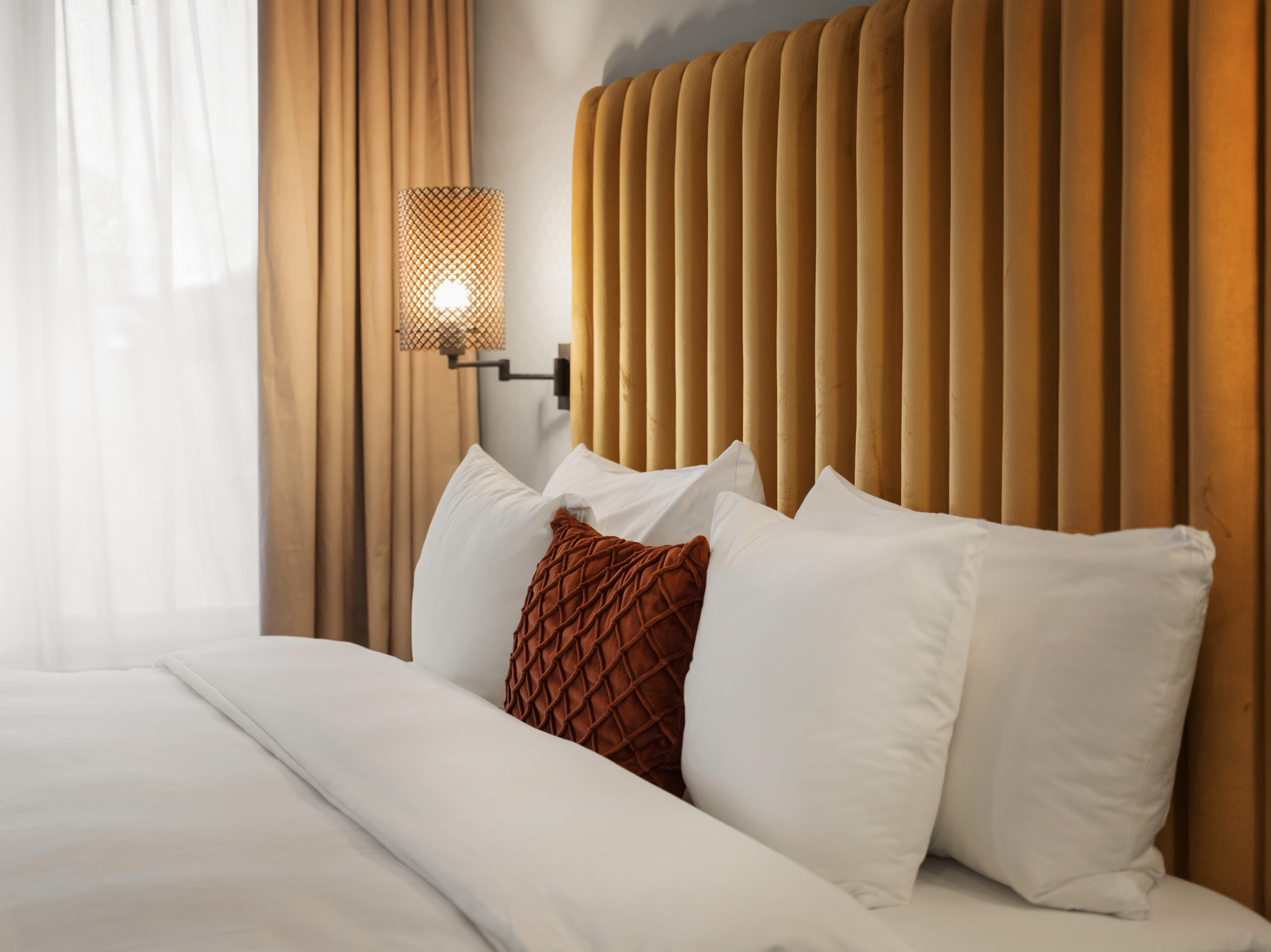 Lyxig hotellrumssäng med senapsgult vadderat huvudgavel och stilfull lampa på väggen, samt vita sängkläder och en dekorativ orange kudde