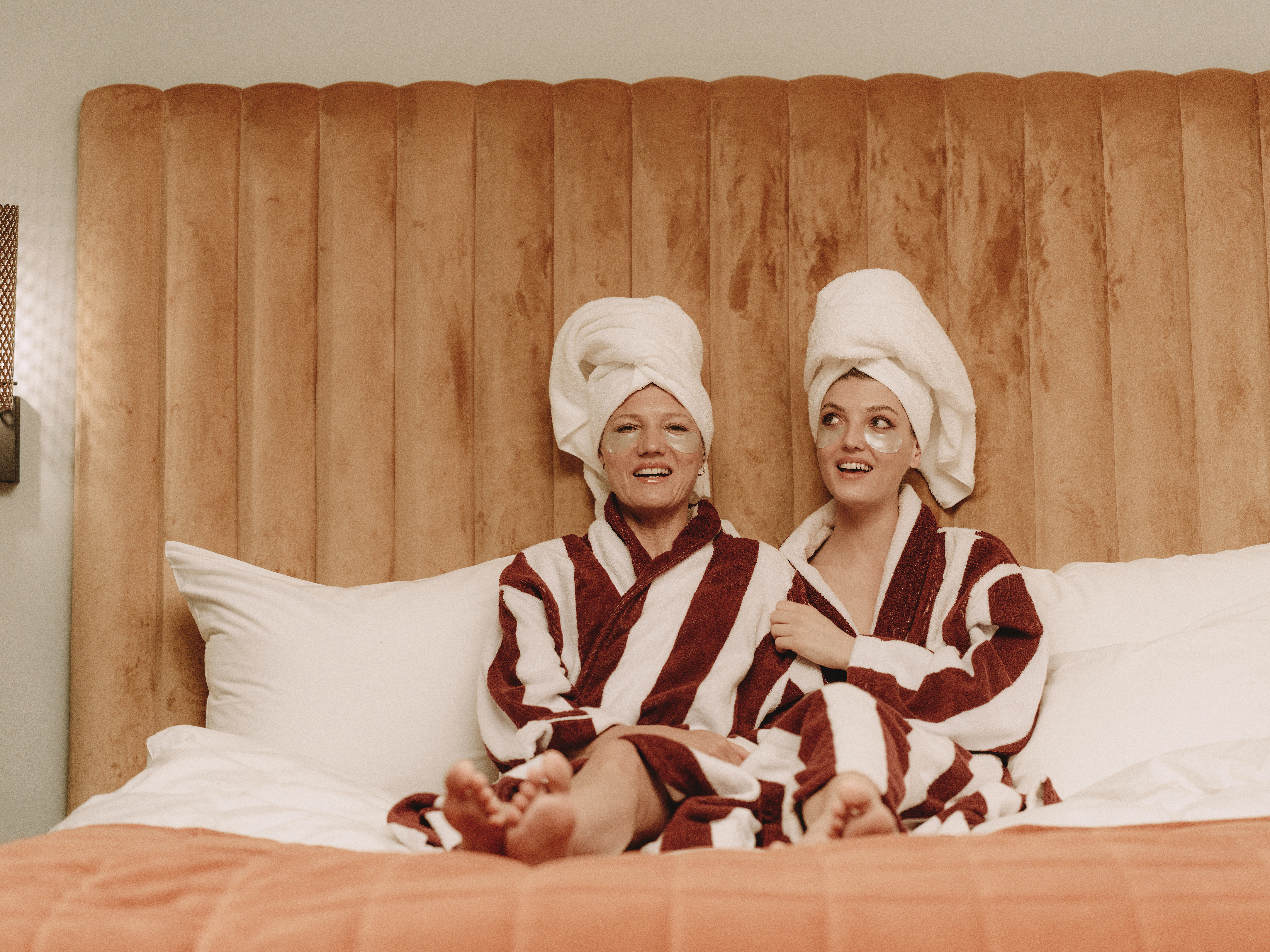 Två personer i randiga badrockar och handdukar på huvudet sitter glada i en hotellsäng