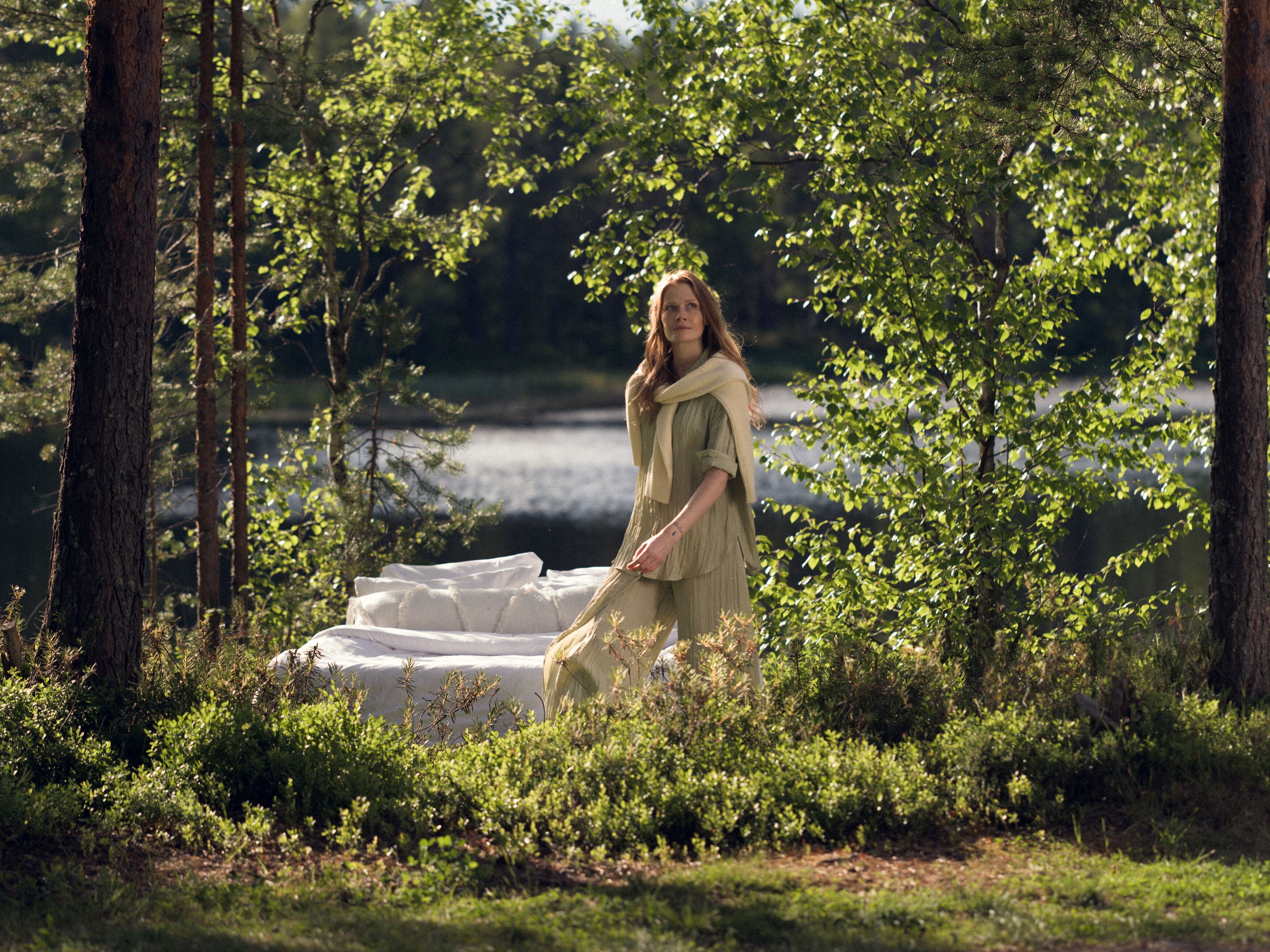 Kvinna i sommarklänning går vid en skogstjärn i skogen, bredvid en vit soffa. Soligt väder och grönskande omgivning.