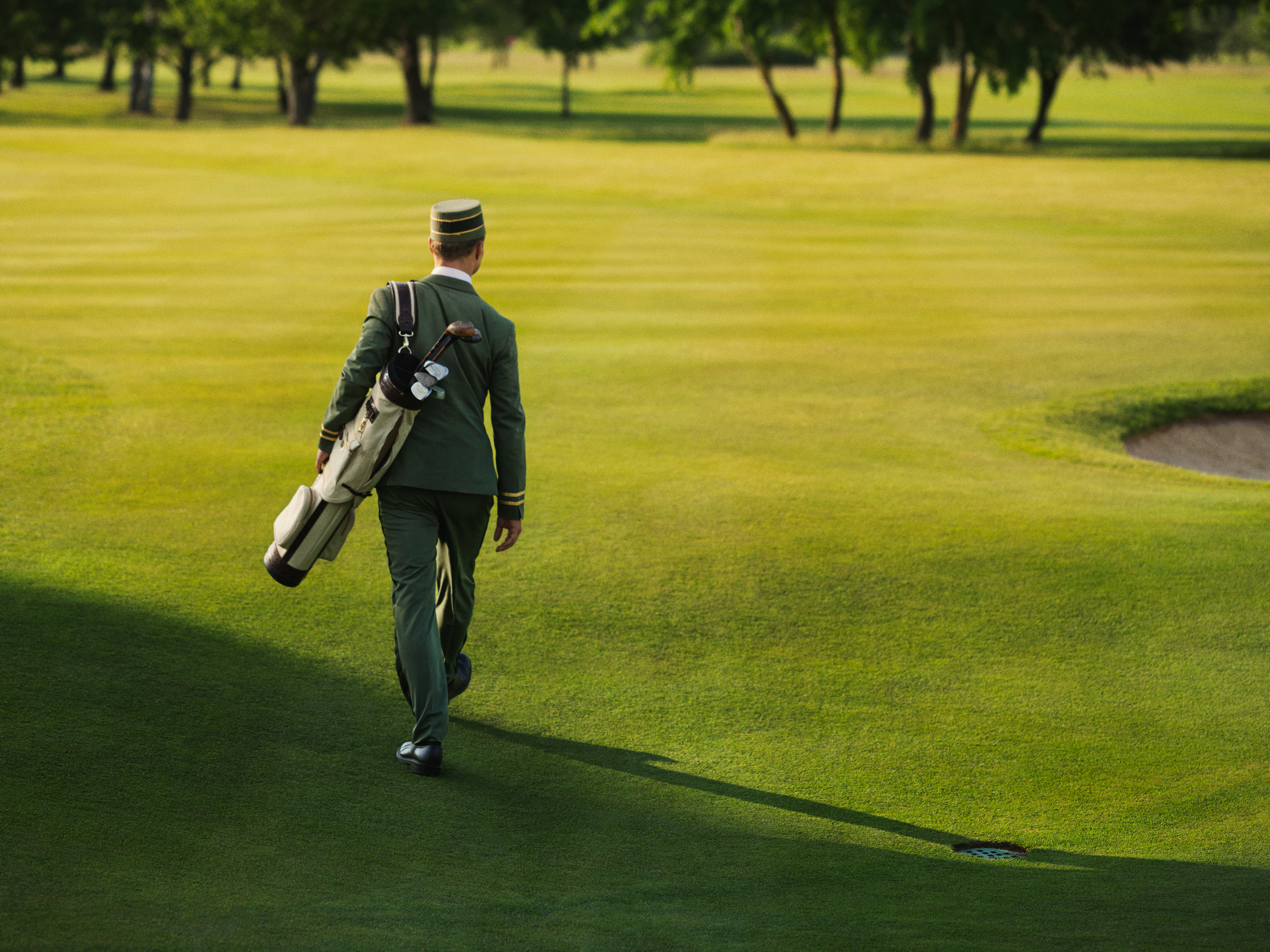 Person in a uniform walking across a sunny golf course, carrying a golf bag with clubs, surrounded by vibrant green grass and trees.