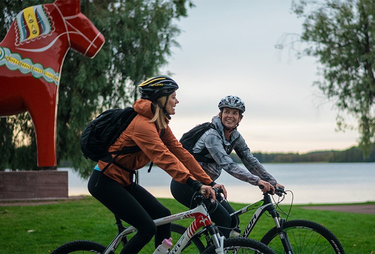 Två glada cyklister och en dalahäst i bakgrunden